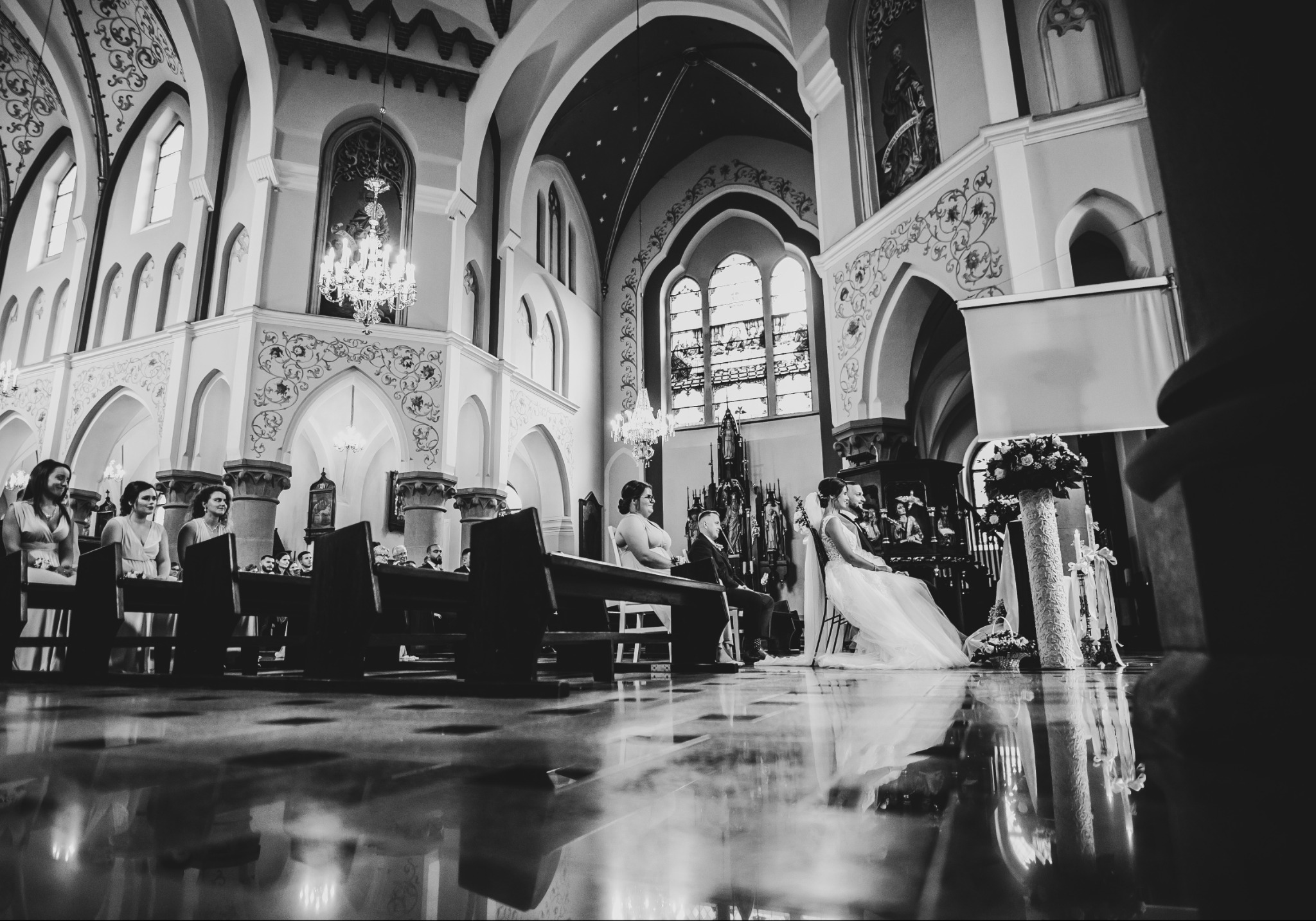 Czarno-białe zdjęcie wnętrza kościoła podczas ceremonii ślubnej, z odbiciem posadzki, ukazujące parę młodą siedzącą na krzesłach przed ołtarzem oraz gości w ławkach.
