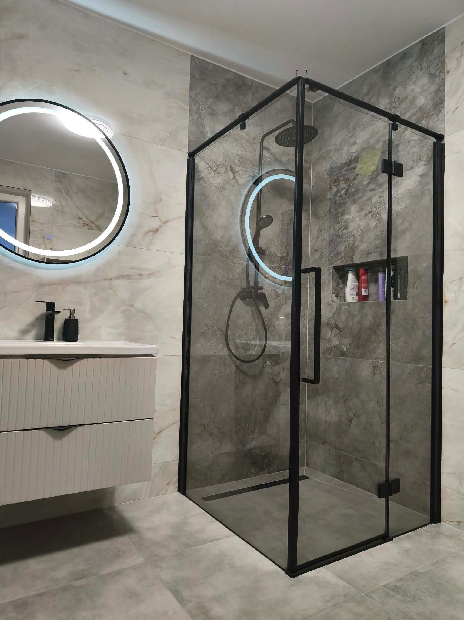 Nowoczesna łazienka z kabiną prysznicową typu walk-in z czarnymi profilami, okrągłym lustrem z podświetleniem LED i szafką pod umywalką w odcieniach szarości.
