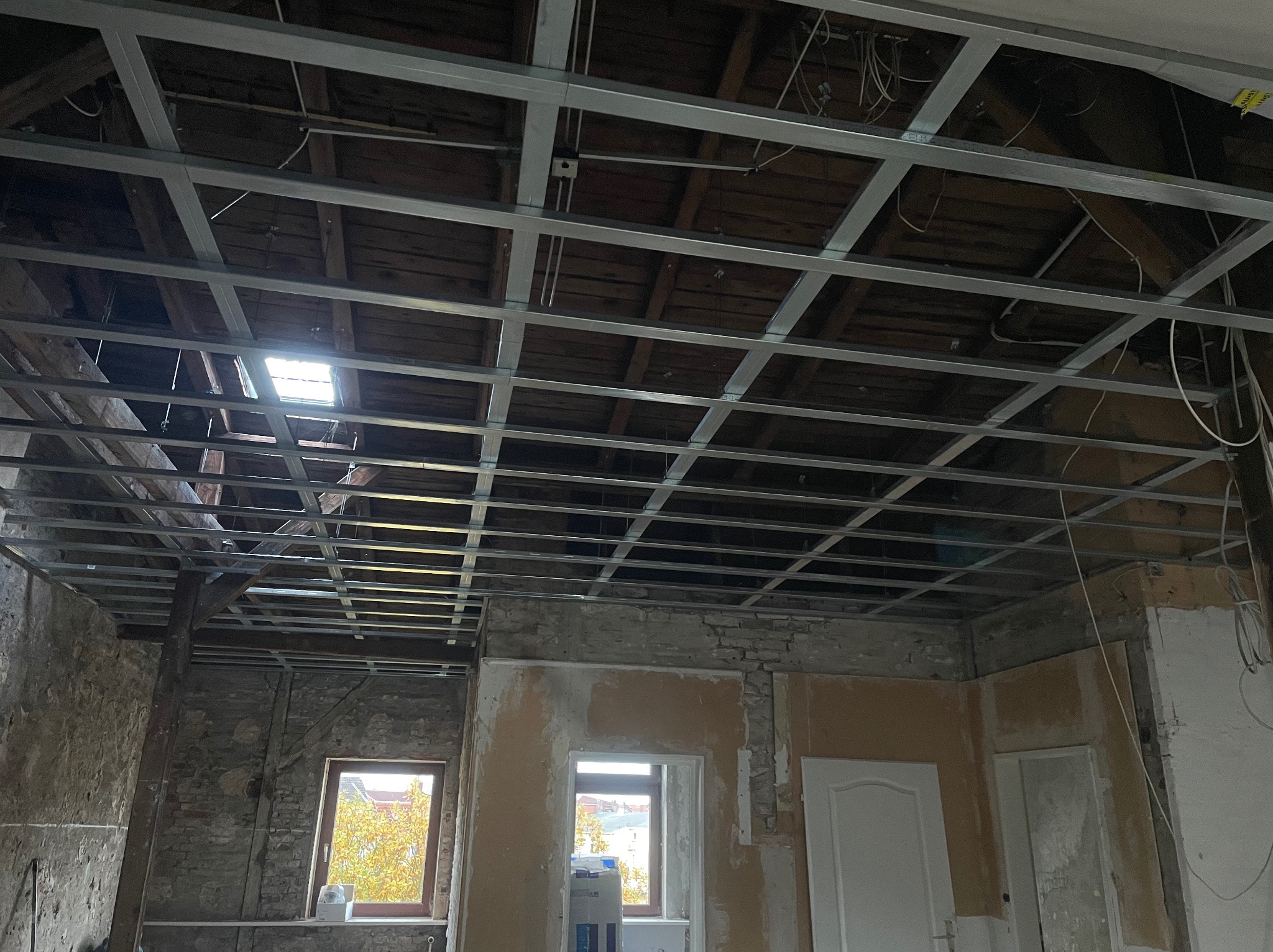 Metalowa konstrukcja sufitu podwieszanego w surowym wnętrzu z oknami i widoczną starą konstrukcją dachu. Prace remontowe w toku, widoczne kable i elementy instalacji.