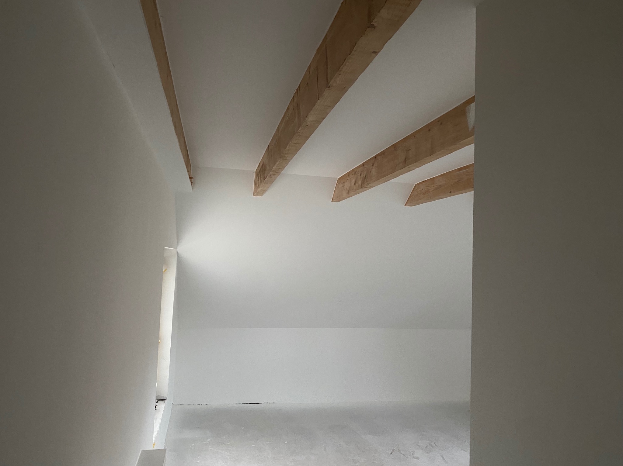 Białe wnętrze poddasza z drewnianymi belkami stropowymi, świeżo po malowaniu. Widoczne detale wykończenia ścian i sufitu, minimalistyczny styl.