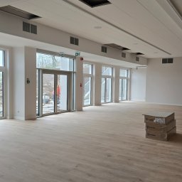 Jasne panele podłogowe w przestronnym, nowoczesnym wnętrzu z dużymi oknami i stosem kartonów z panelami w rogu. Widoczne elementy wentylacji pod sufitem.