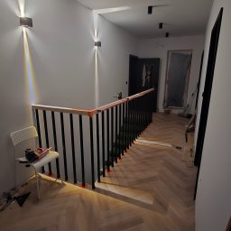 Ułożona podłoga z paneli w jodełkę francuską na korytarzu, widoczne narzędzia i krzesło, zamontowane oświetlenie ścienne, czarne balustrady schodowe.