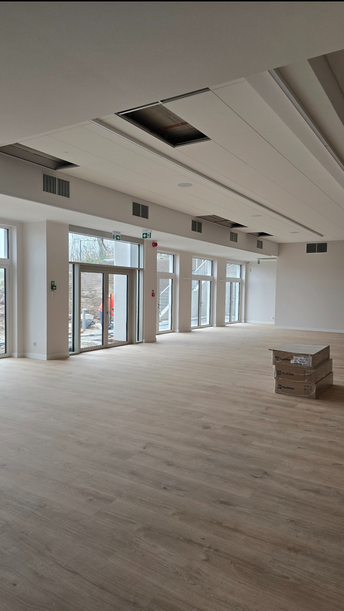 Jasne panele podłogowe w przestronnym, nowoczesnym wnętrzu z dużymi oknami i stosem kartonów z panelami w rogu. Widoczne elementy wentylacji pod sufitem.