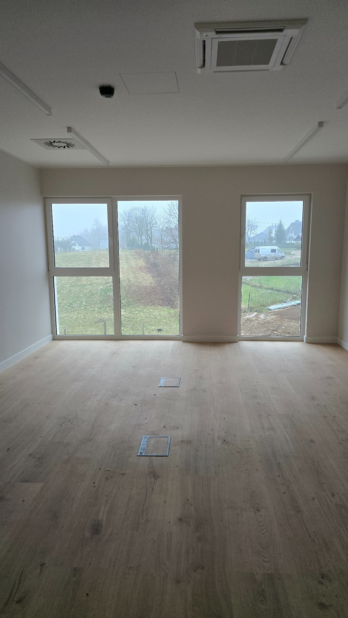 Jasne panele podłogowe w pustym pokoju z dwoma oknami. Widoczne kratki wentylacyjne w podłodze i na suficie. Minimalistyczne wnętrze w neutralnej kolorystyce.