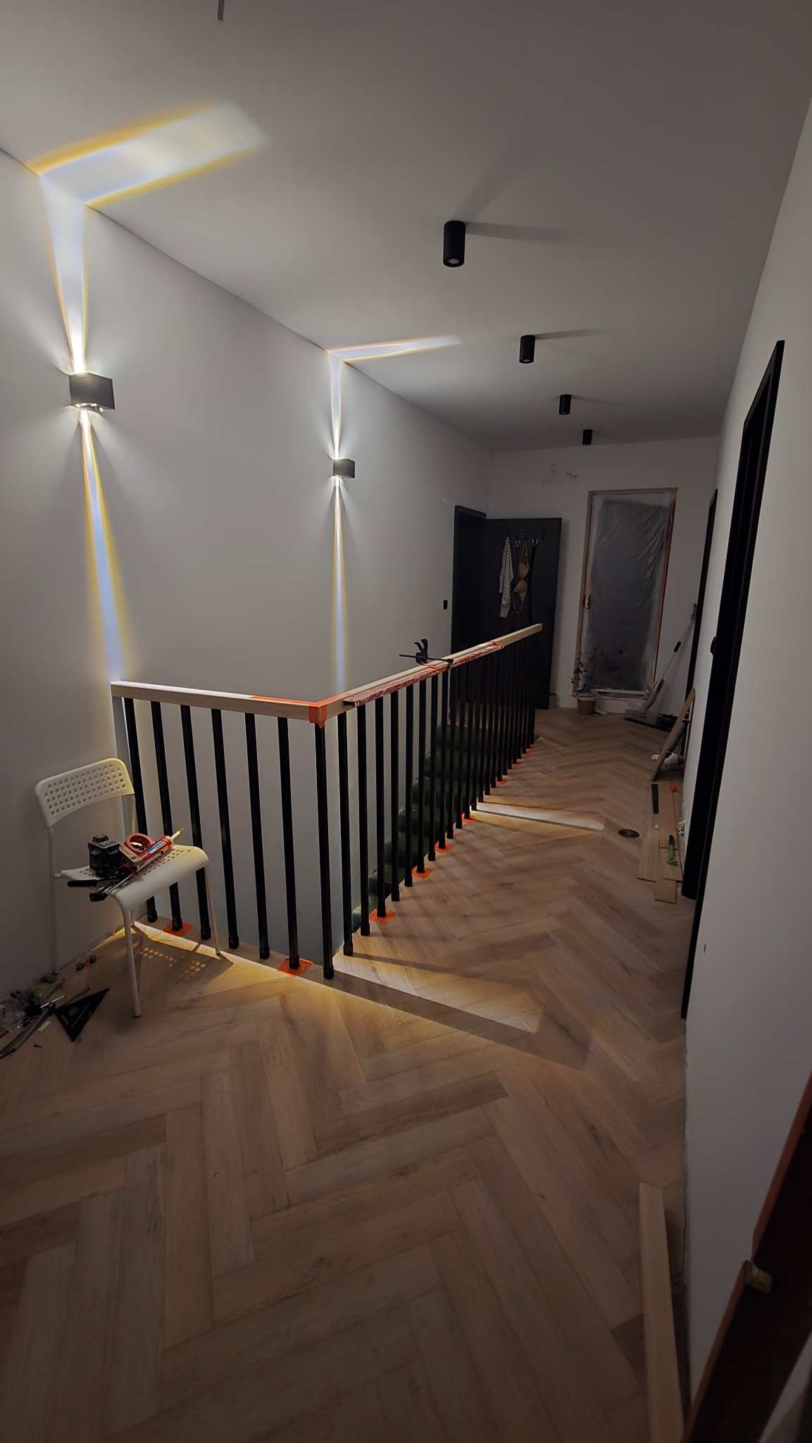 Ułożona podłoga z paneli w jodełkę francuską na korytarzu, widoczne narzędzia i krzesło, zamontowane oświetlenie ścienne, czarne balustrady schodowe.