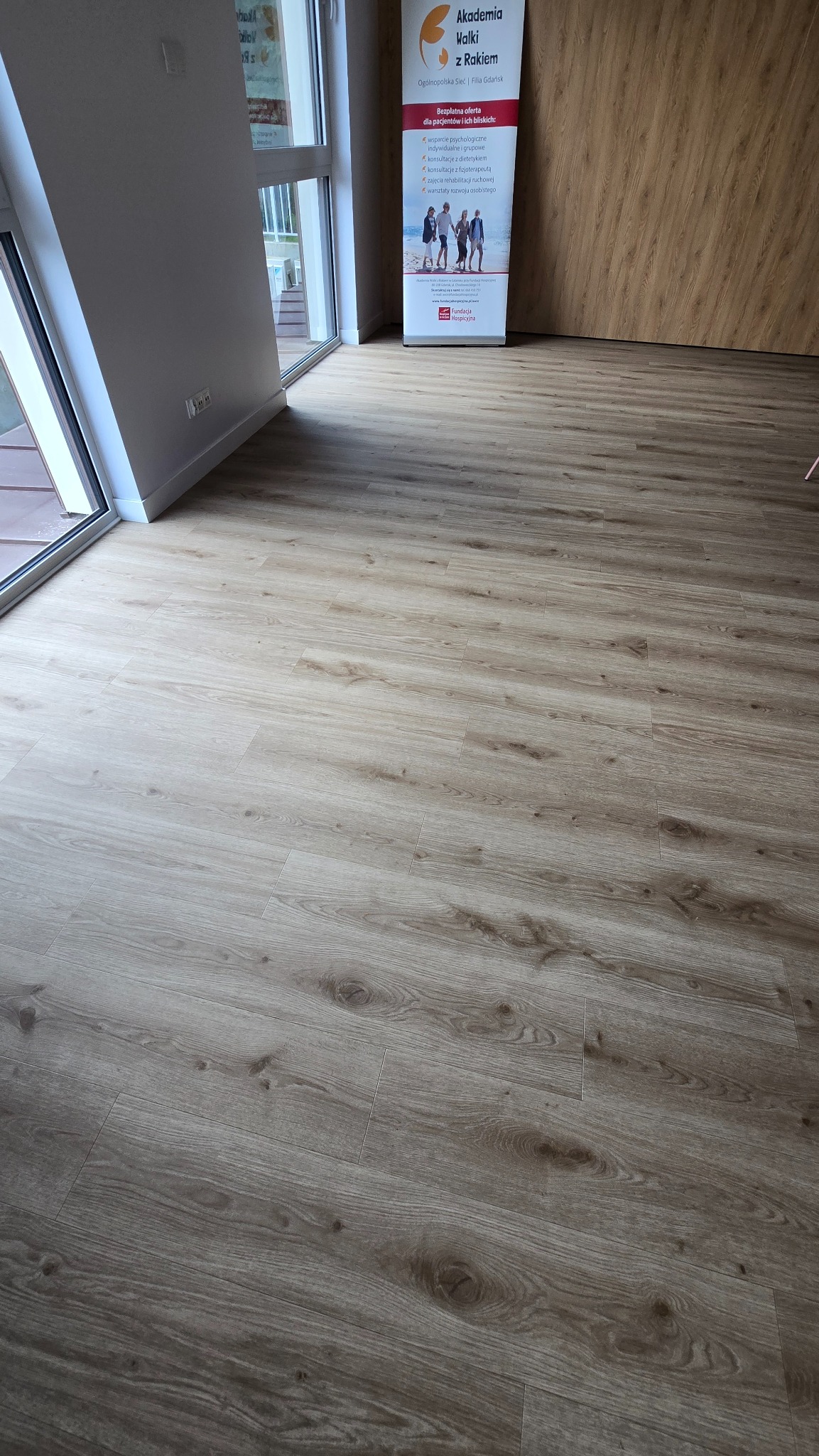 Jasne panele podłogowe z widoczną strukturą drewna, ułożone w nowym pomieszczeniu. W tle okno i baner reklamowy.