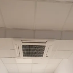 Kasetonowa jednostka klimatyzacyjna zamontowana w suficie podwieszanym, widok z dołu, w tle widoczny znak ewakuacyjny.