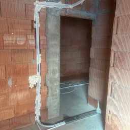 Futuro - Mateusz Sawicki - Montaż instalacji klimatyzacji w surowym wnętrzu: białe rurki prowadzone wzdłuż ściany z cegieł, zabezpieczone pianką montażową przy framudze drzwi.