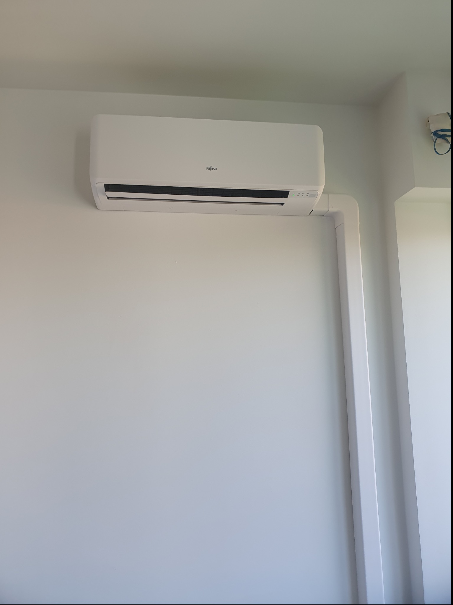 Biała jednostka klimatyzacyjna Fujitsu zamontowana na białej ścianie, z widocznymi elementami instalacji w białej osłonie.