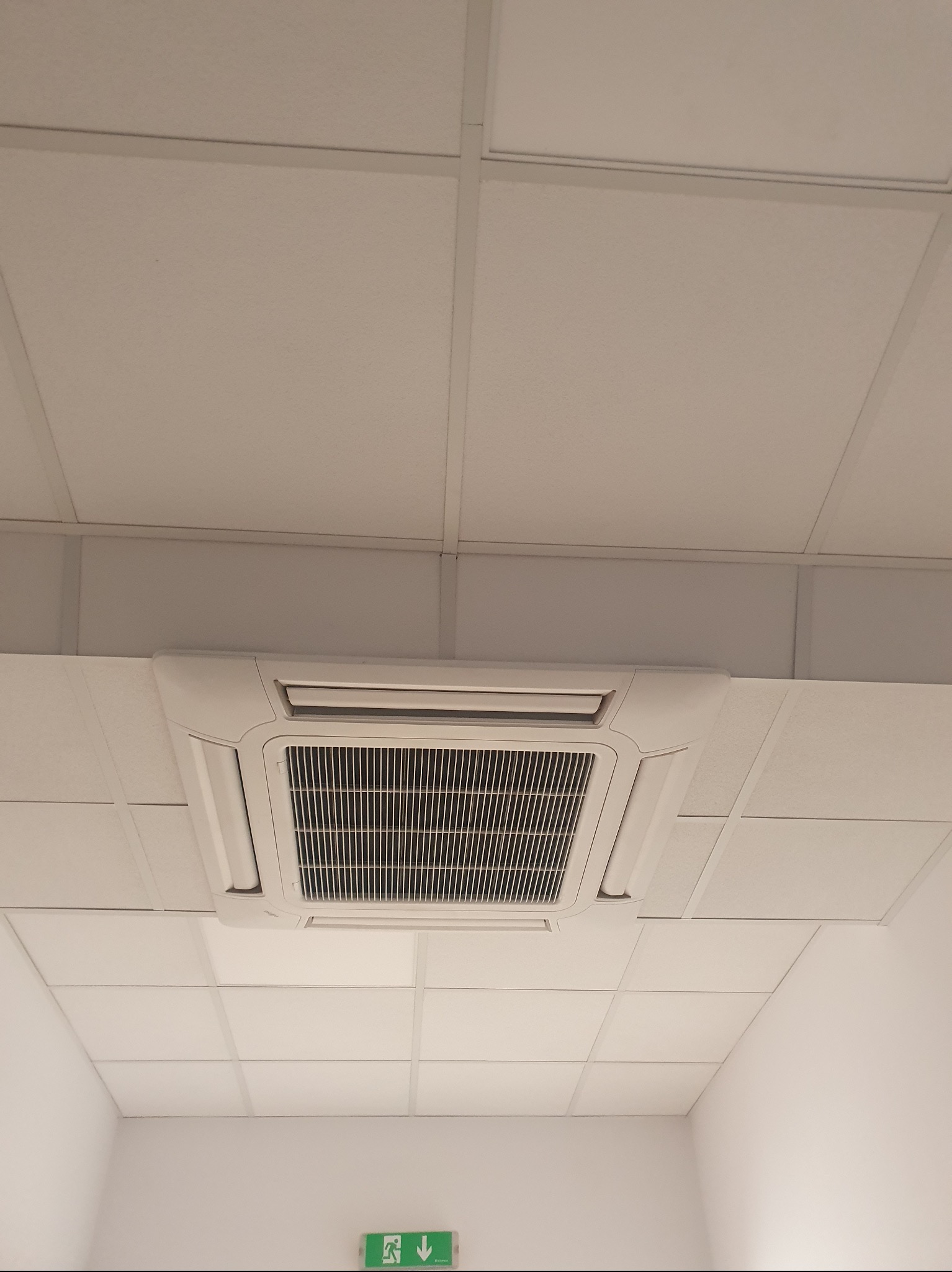 Kasetonowa jednostka klimatyzacyjna zamontowana w suficie podwieszanym, widok z dołu, w tle widoczny znak ewakuacyjny.