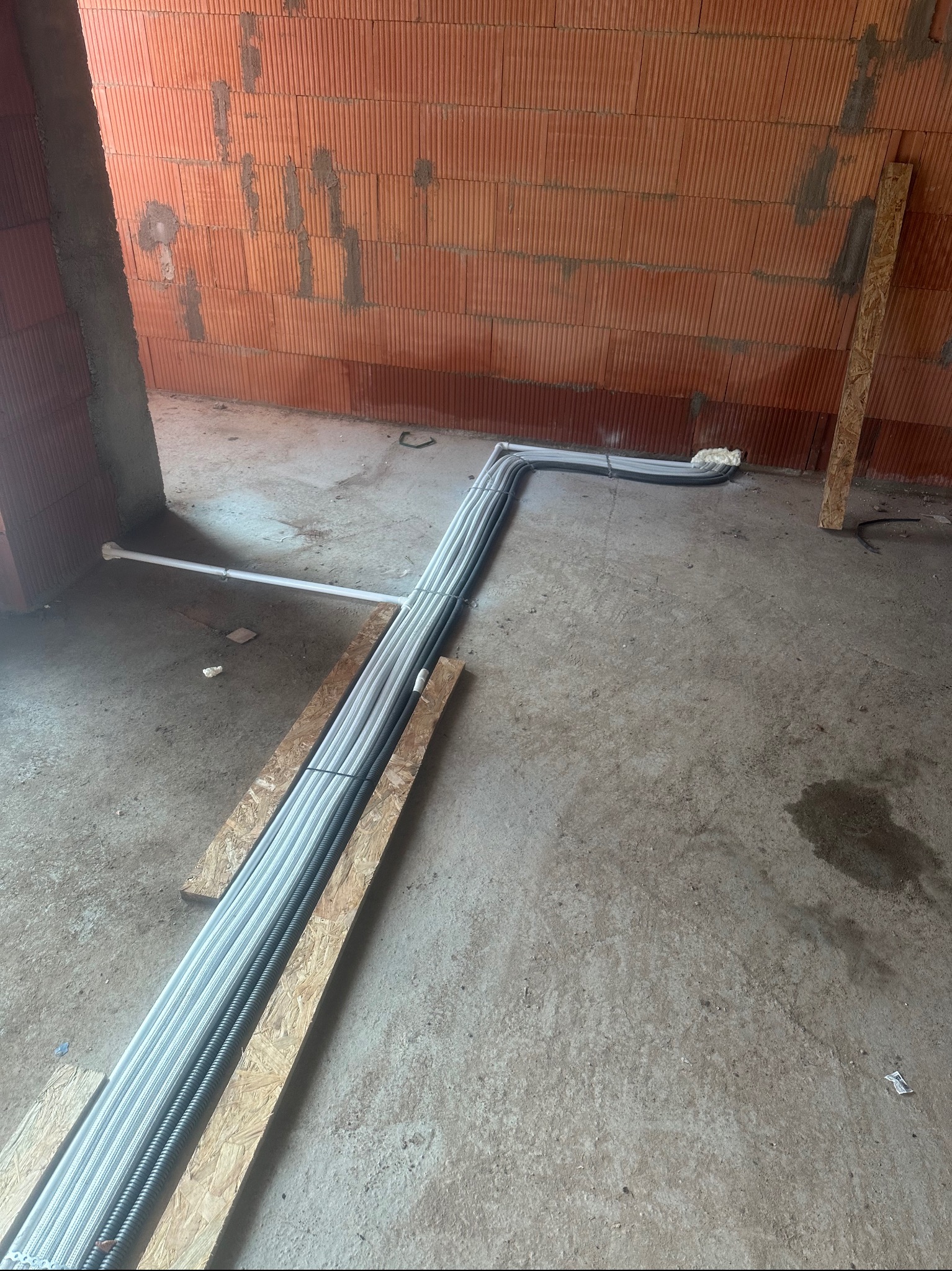 Rury instalacyjne ułożone na deskach w surowym wnętrzu z ceglaną ścianą, widoczne elementy mocowań i podłączeń.