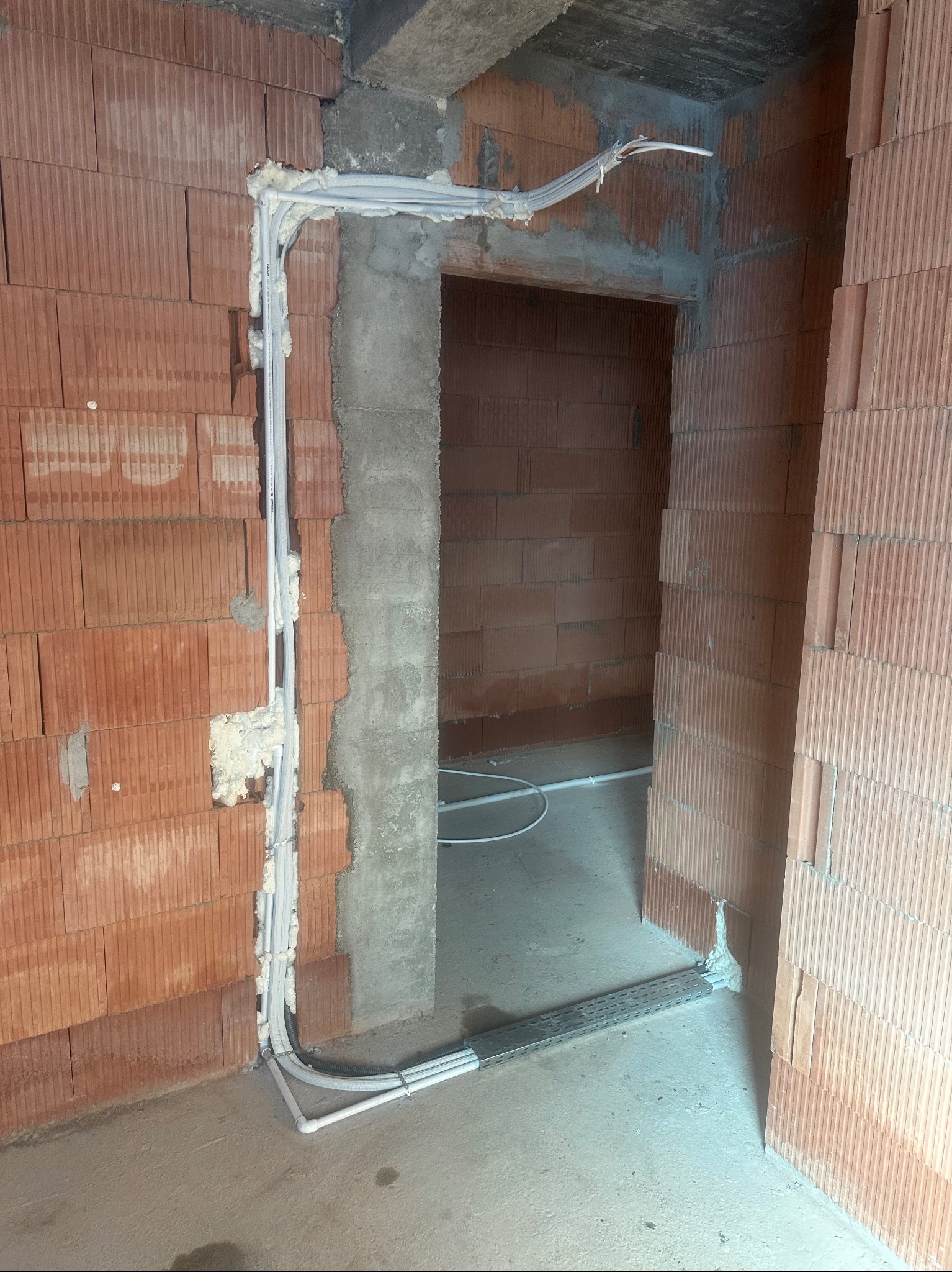 Montaż instalacji klimatyzacji w surowym wnętrzu: białe rurki prowadzone wzdłuż ściany z cegieł, zabezpieczone pianką montażową przy framudze drzwi.