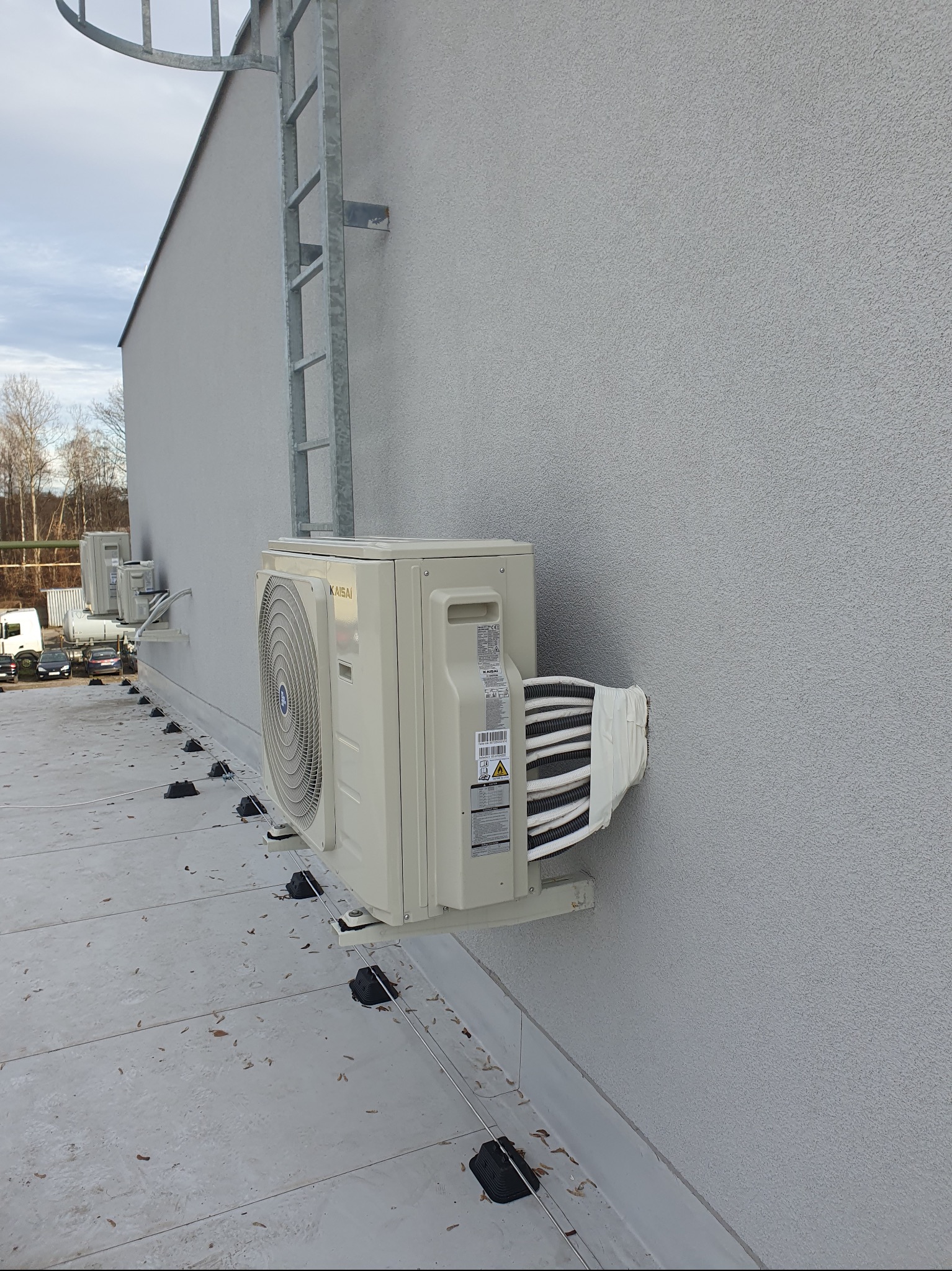 Zewnętrzna jednostka klimatyzacji Kaisai zamontowana na szarej elewacji budynku z widoczną drabiną przeciwpożarową i elementami instalacji na dachu.