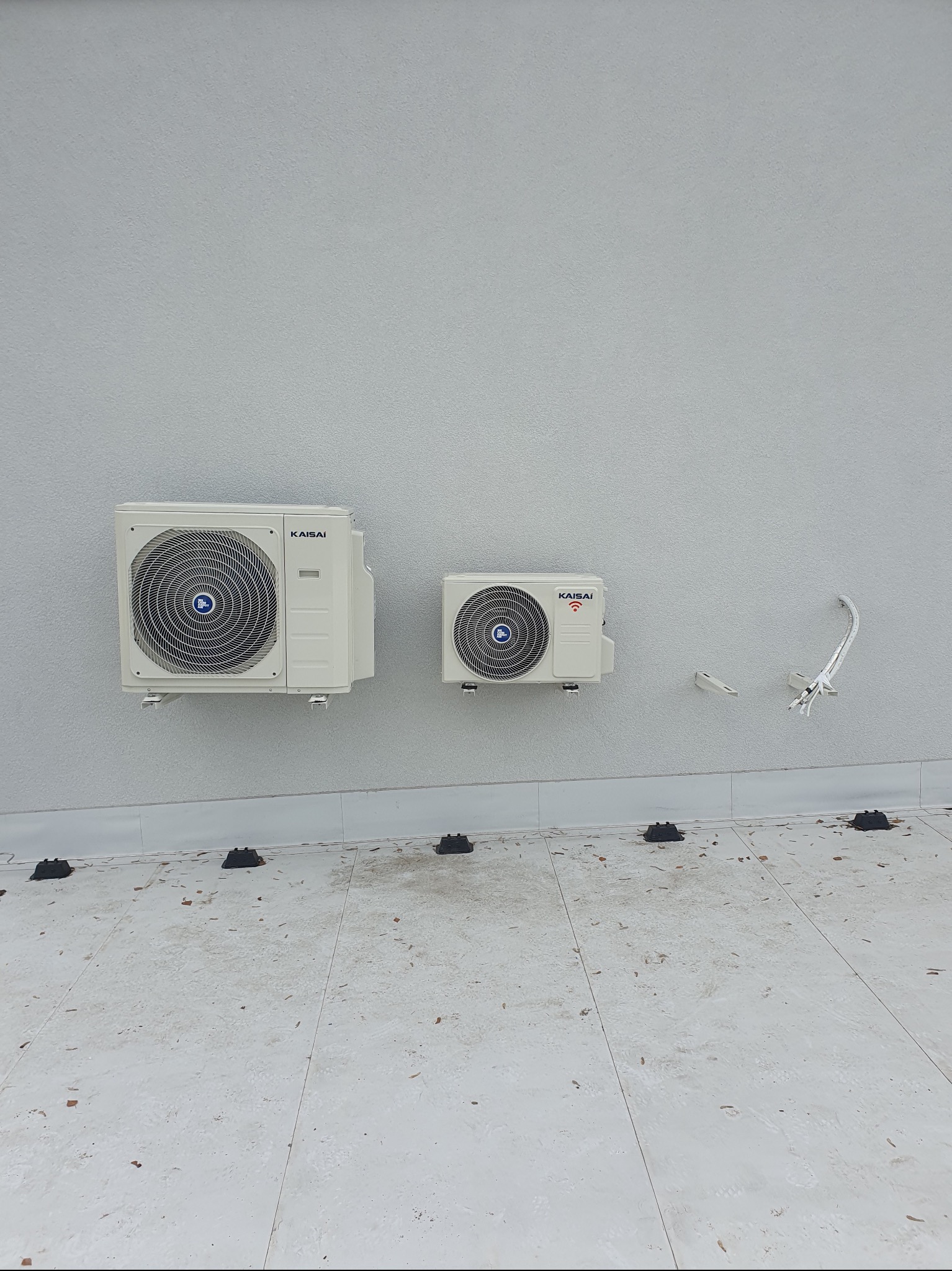 Dwa zamontowane na zewnątrz klimatyzatory marki Kaisai na szarej ścianie, widoczne przewody instalacyjne i czarne podkładki pod agregatami.