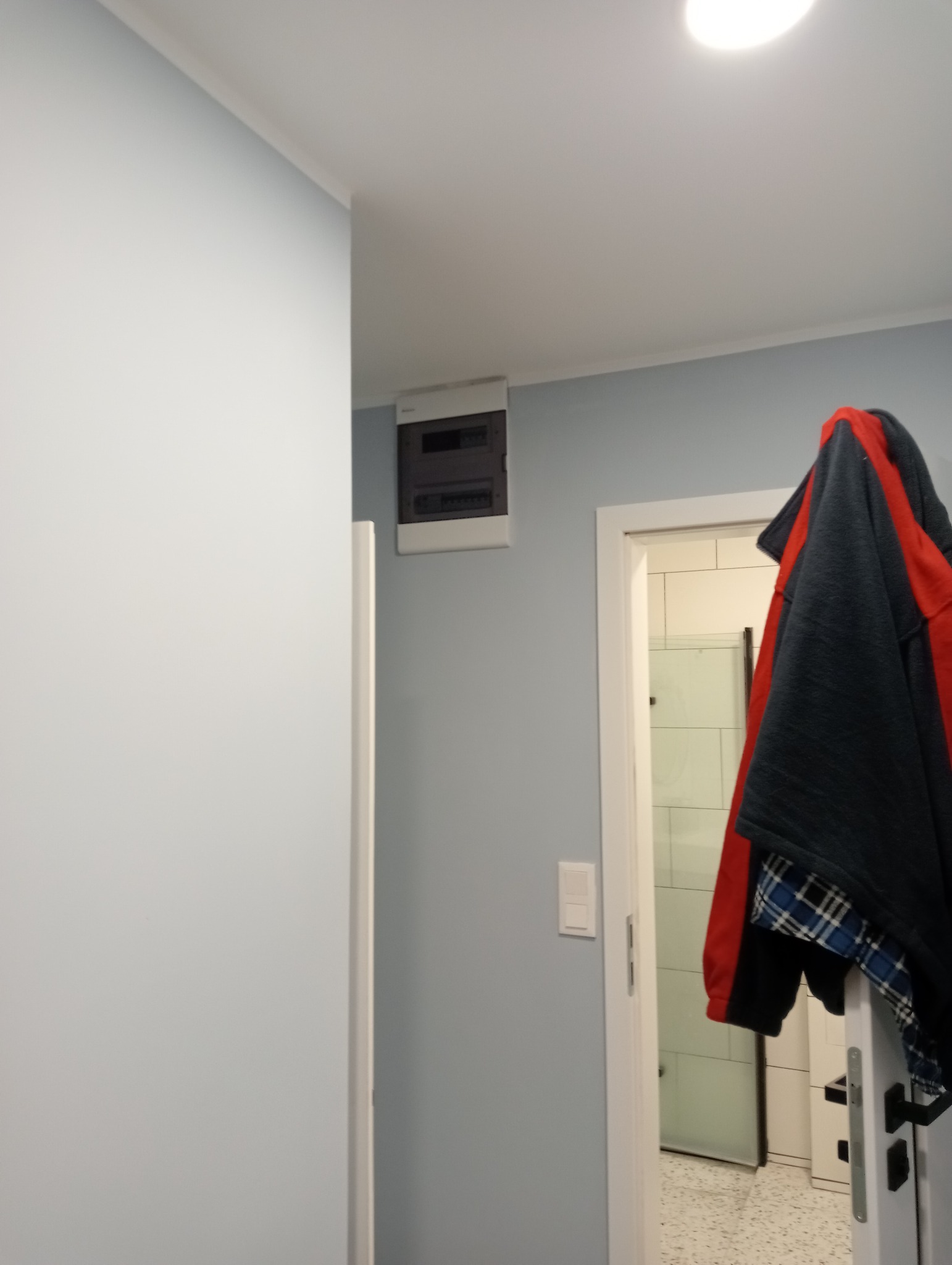 Szara ściana z zamontowaną skrzynką elektryczną i włącznikiem światła, widok na łazienkę przez otwarte drzwi, ubrania wiszące na drzwiach.
