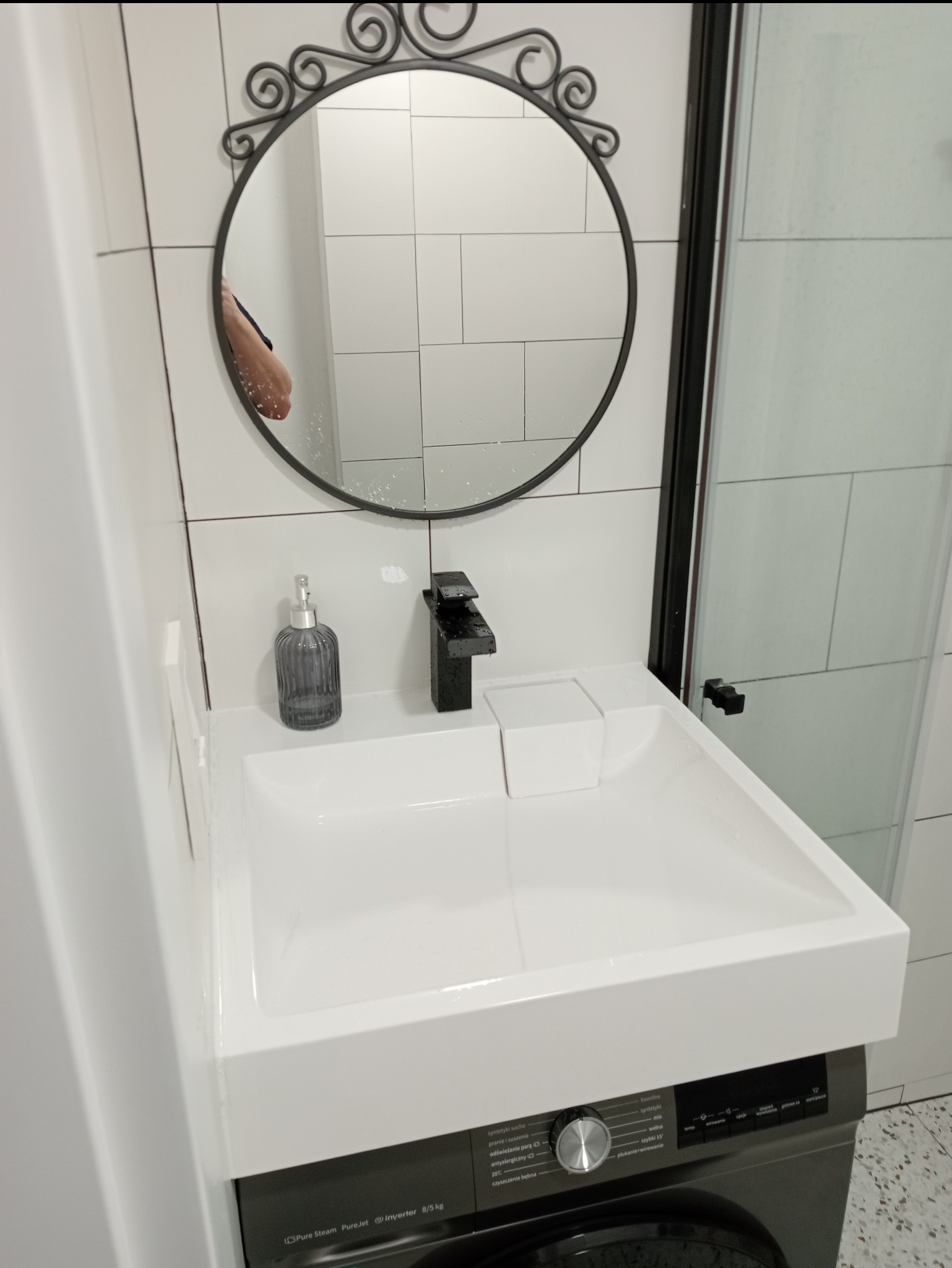 Nowoczesna łazienka z białą umywalką nablatową, czarną armaturą i dekoracyjnym okrągłym lustrem nad pralką automatyczną.