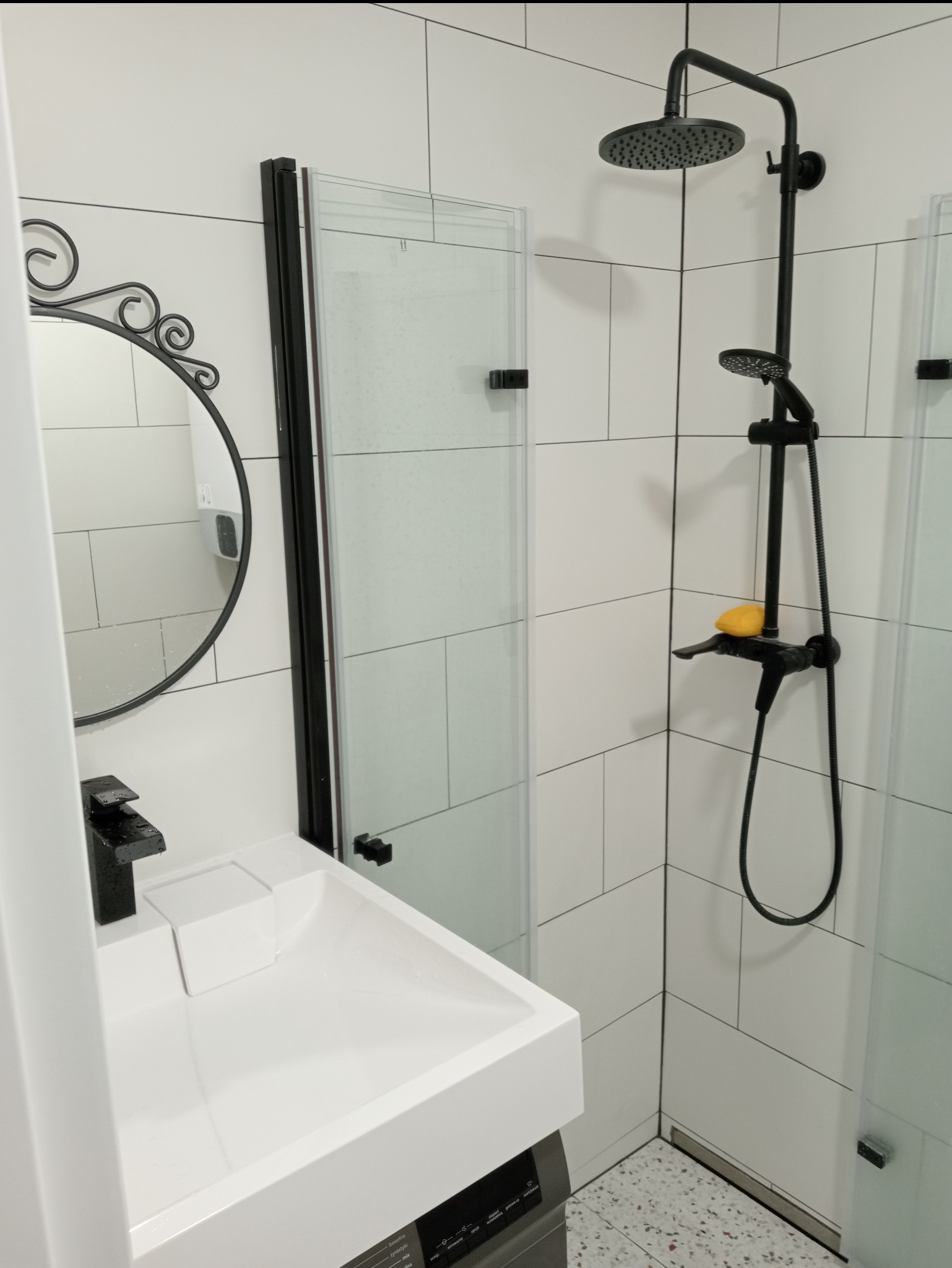 Nowoczesna łazienka z czarną armaturą: deszczownica, bateria umywalkowa, okrągłe lustro z ozdobnym obramowaniem i białe płytki imitujące cegłę.