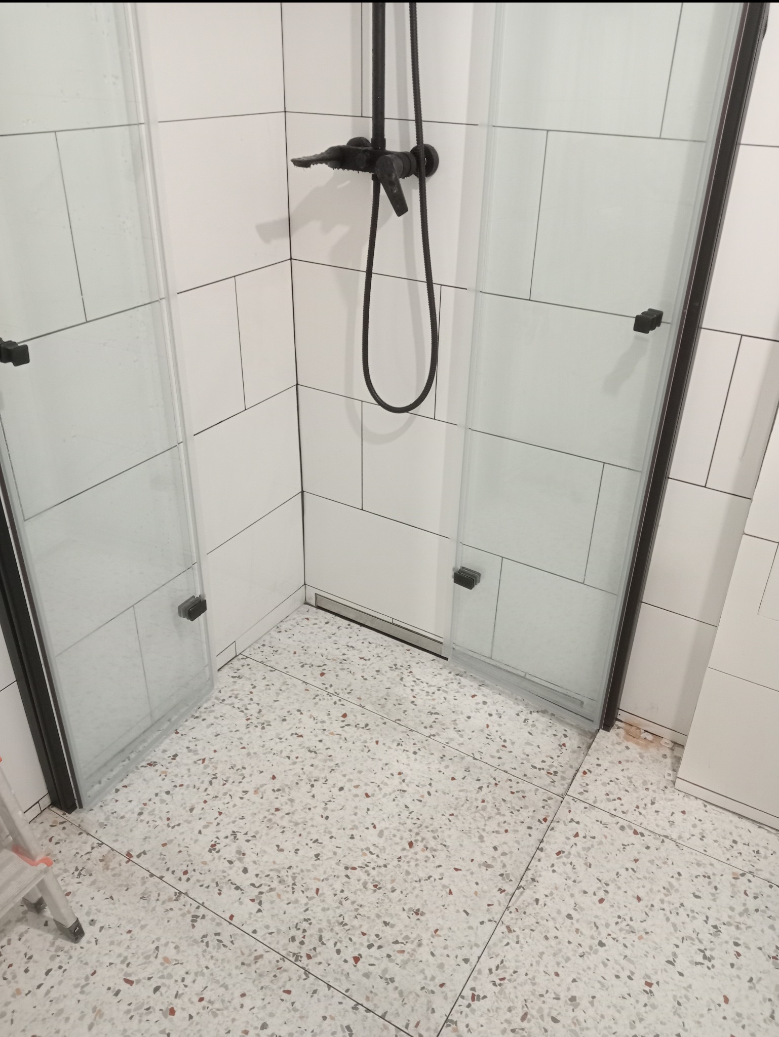 Nowoczesna kabina prysznicowa z czarną armaturą i białymi płytkami ściennymi, posadzka z lastryko, widoczny odpływ liniowy.