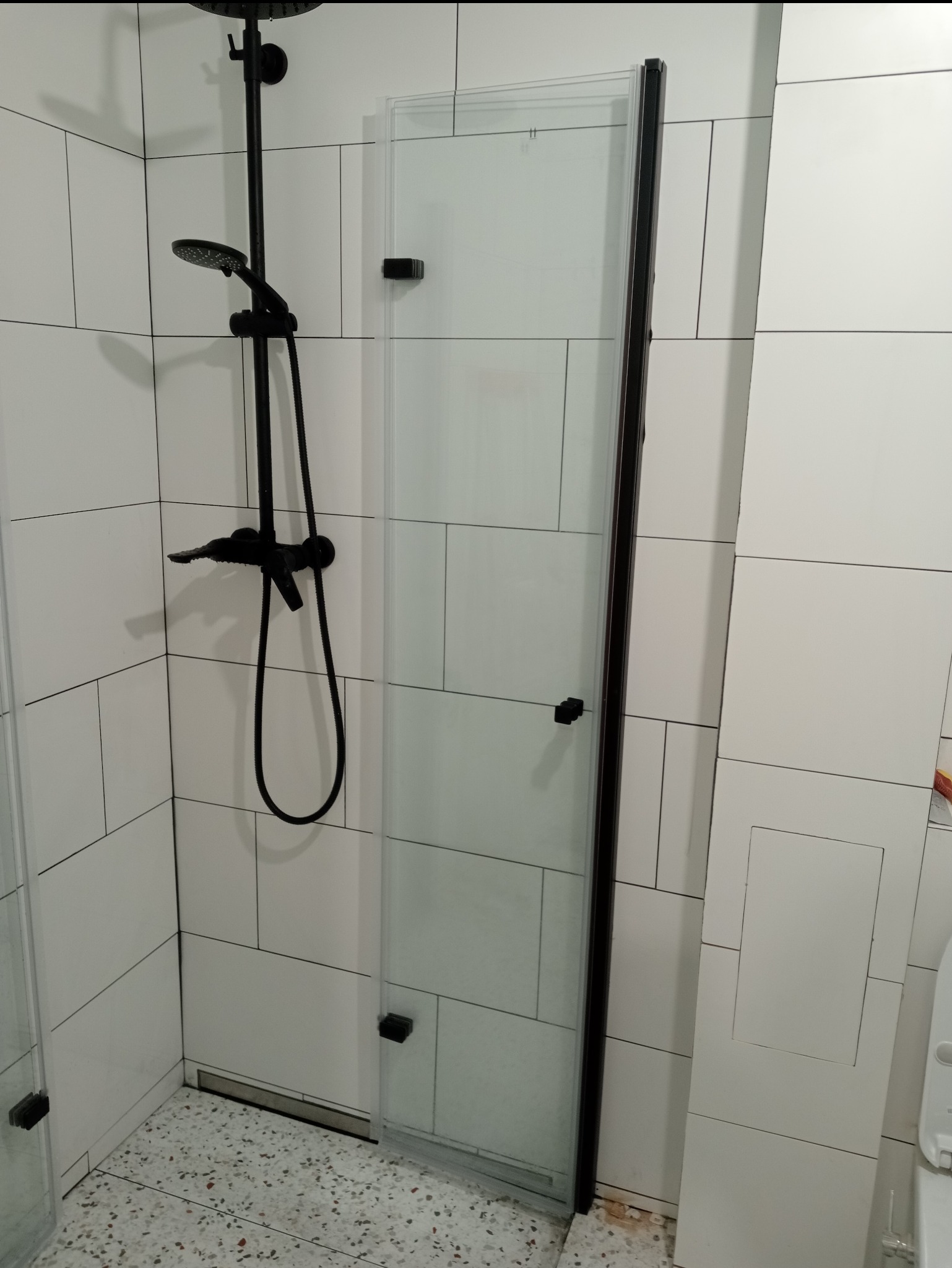 Zamontowana szklana kabina prysznicowa z czarnymi profilami i armaturą w łazience wyłożonej białymi, prostokątnymi płytkami.