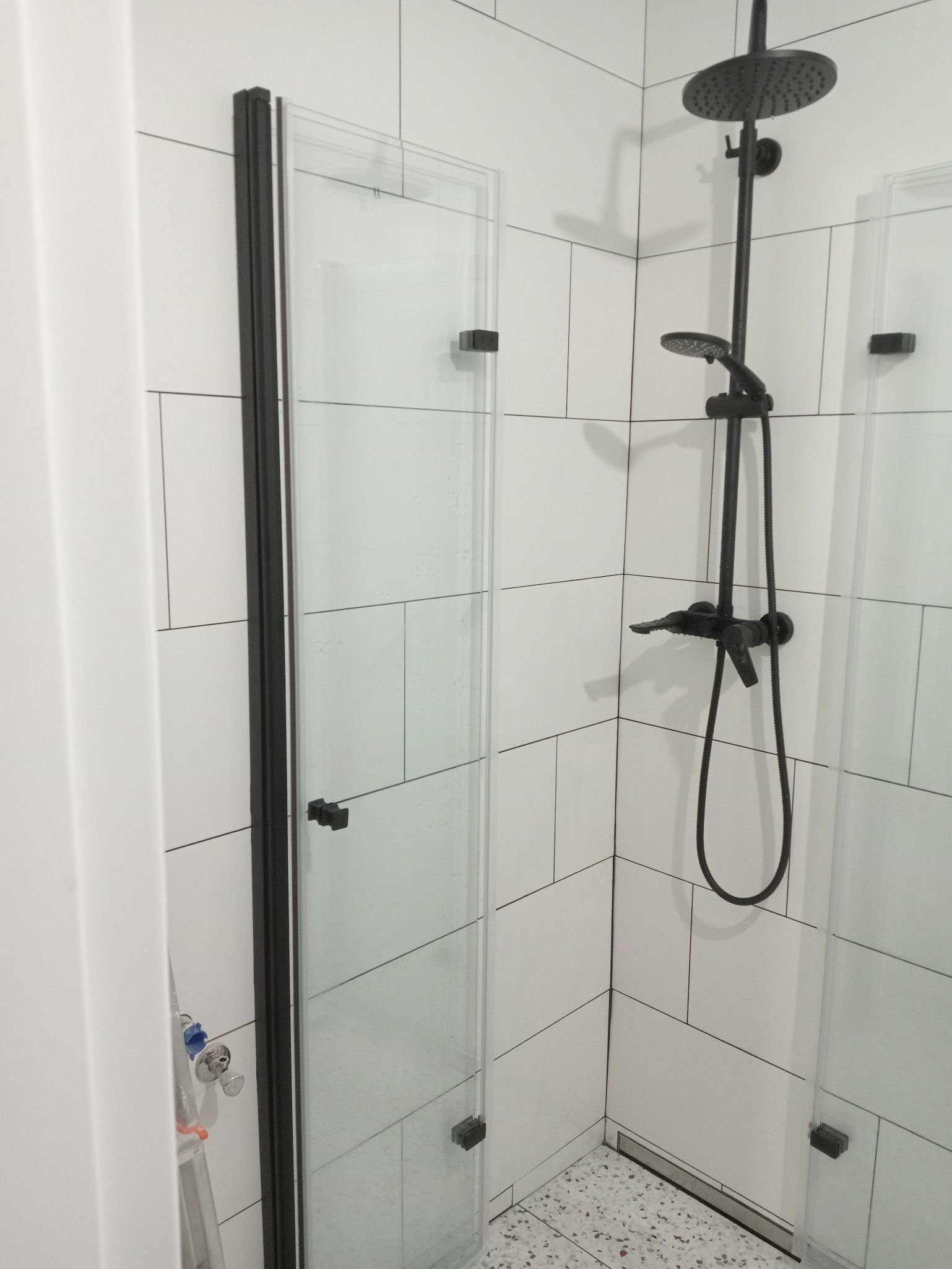 Nowoczesna kabina prysznicowa z czarną armaturą i białymi, prostokątnymi płytkami na ścianach, widoczny fragment drabiny malarskiej z niebieskim zaworem po lewej stronie.