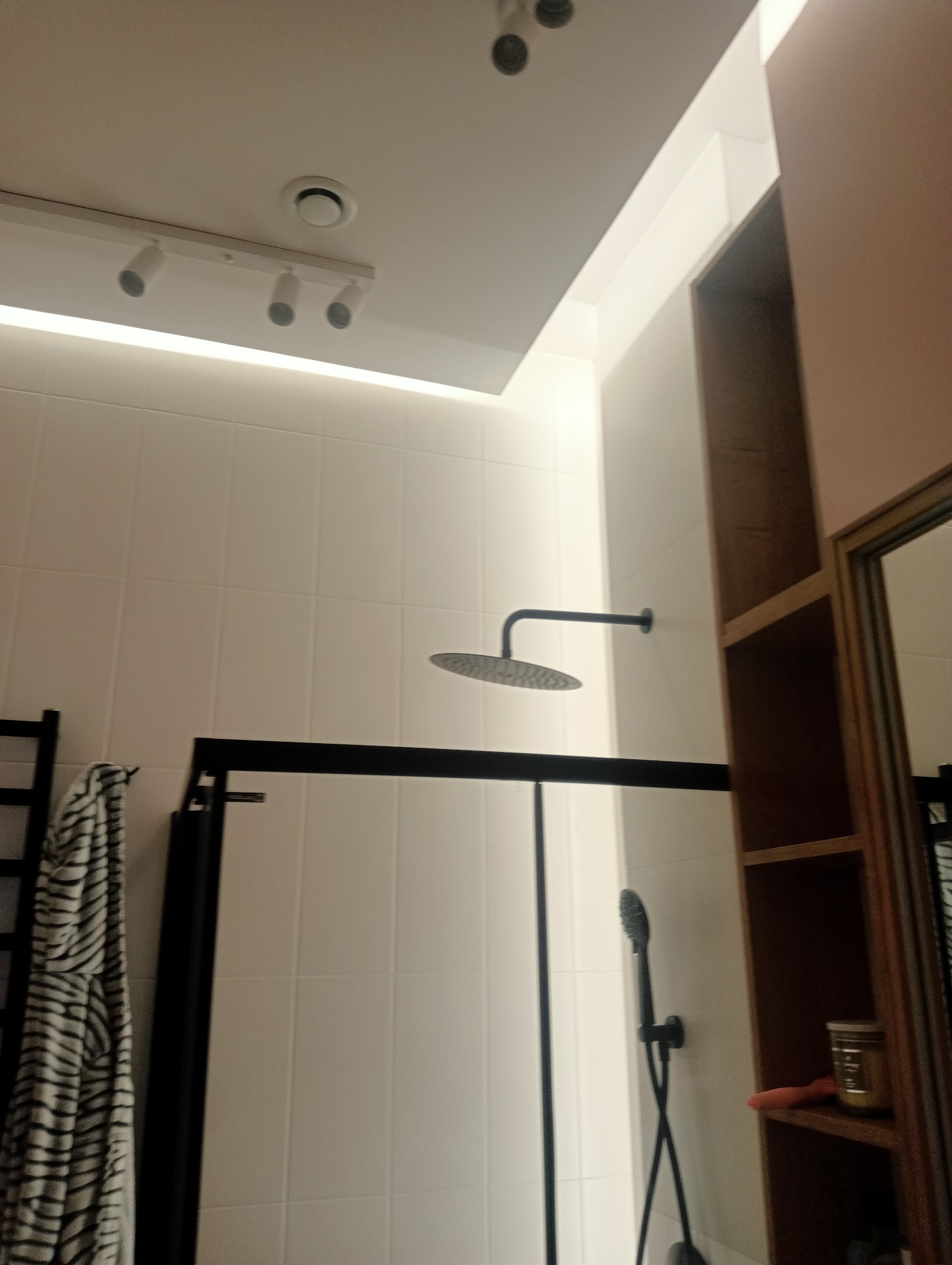 Nowoczesna kabina prysznicowa walk-in z czarną armaturą, deszczownicą i panelem prysznicowym na tle białych płytek, oświetlenie punktowe i LED, widoczny fragment regału z akcesoriami.