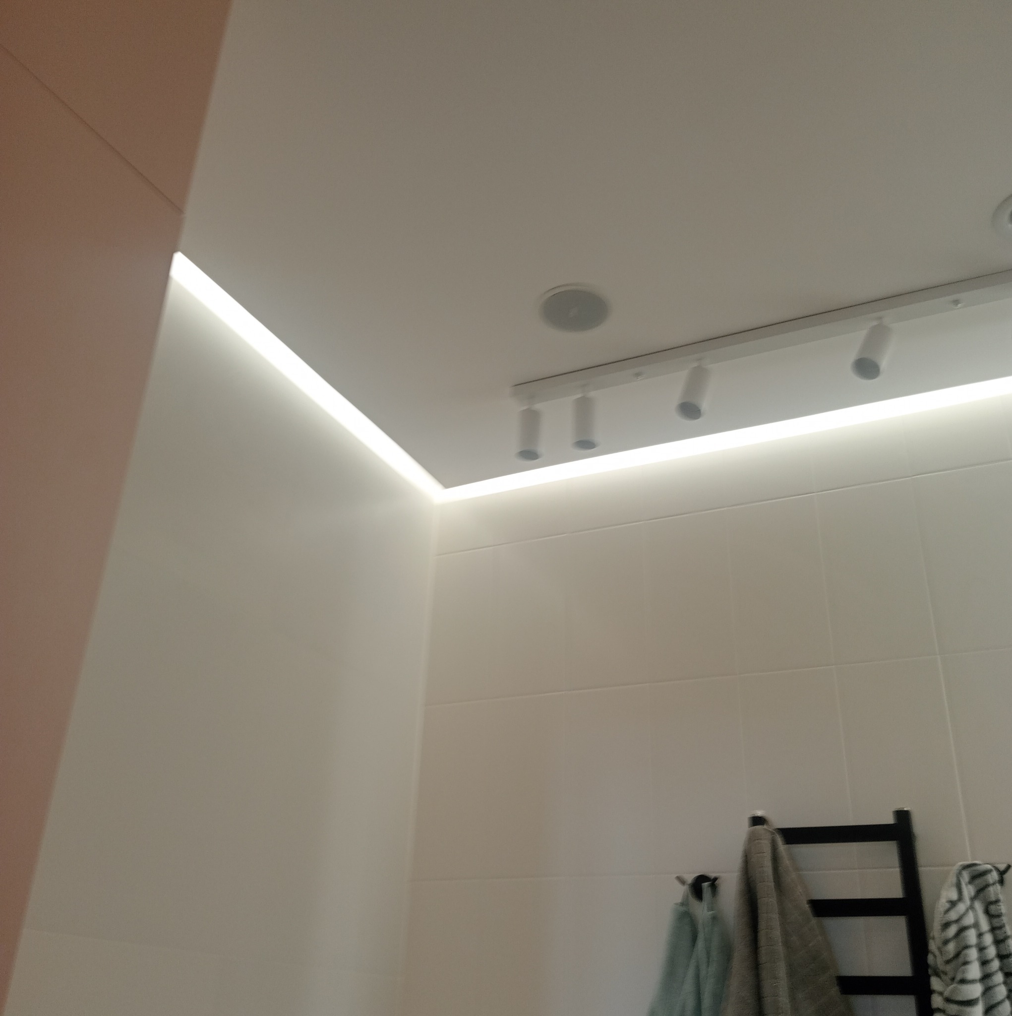 Nowoczesna łazienka z podwieszanym sufitem, oświetleniem LED wzdłuż krawędzi i reflektorami punktowymi na szynie, widoczny fragment grzejnika drabinkowego z ręcznikami.