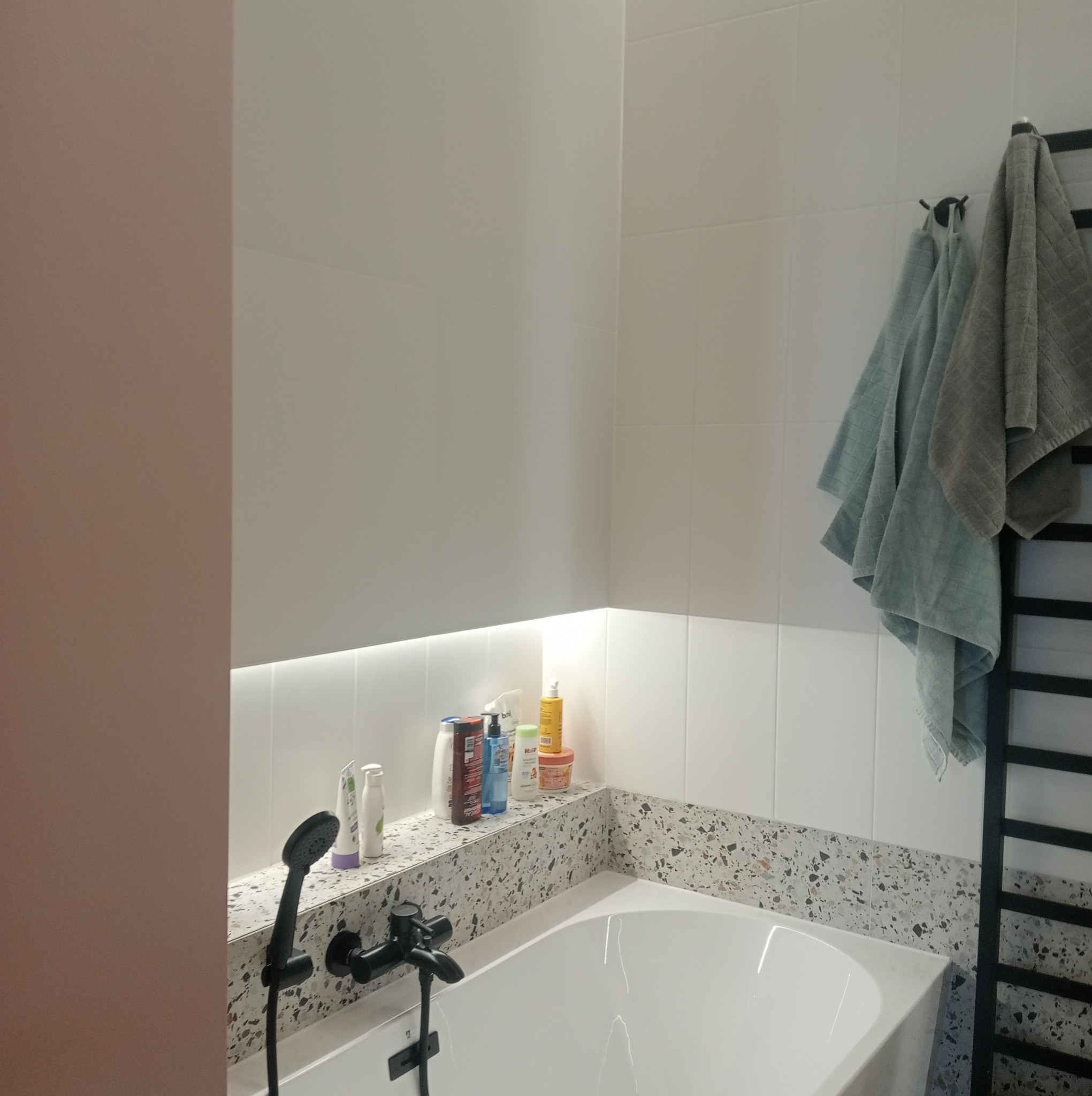 Nowoczesna łazienka z białą wanną, czarną armaturą i półką z terazzo podświetloną ledami. Na półce kosmetyki, na grzejniku wiszą ręczniki.