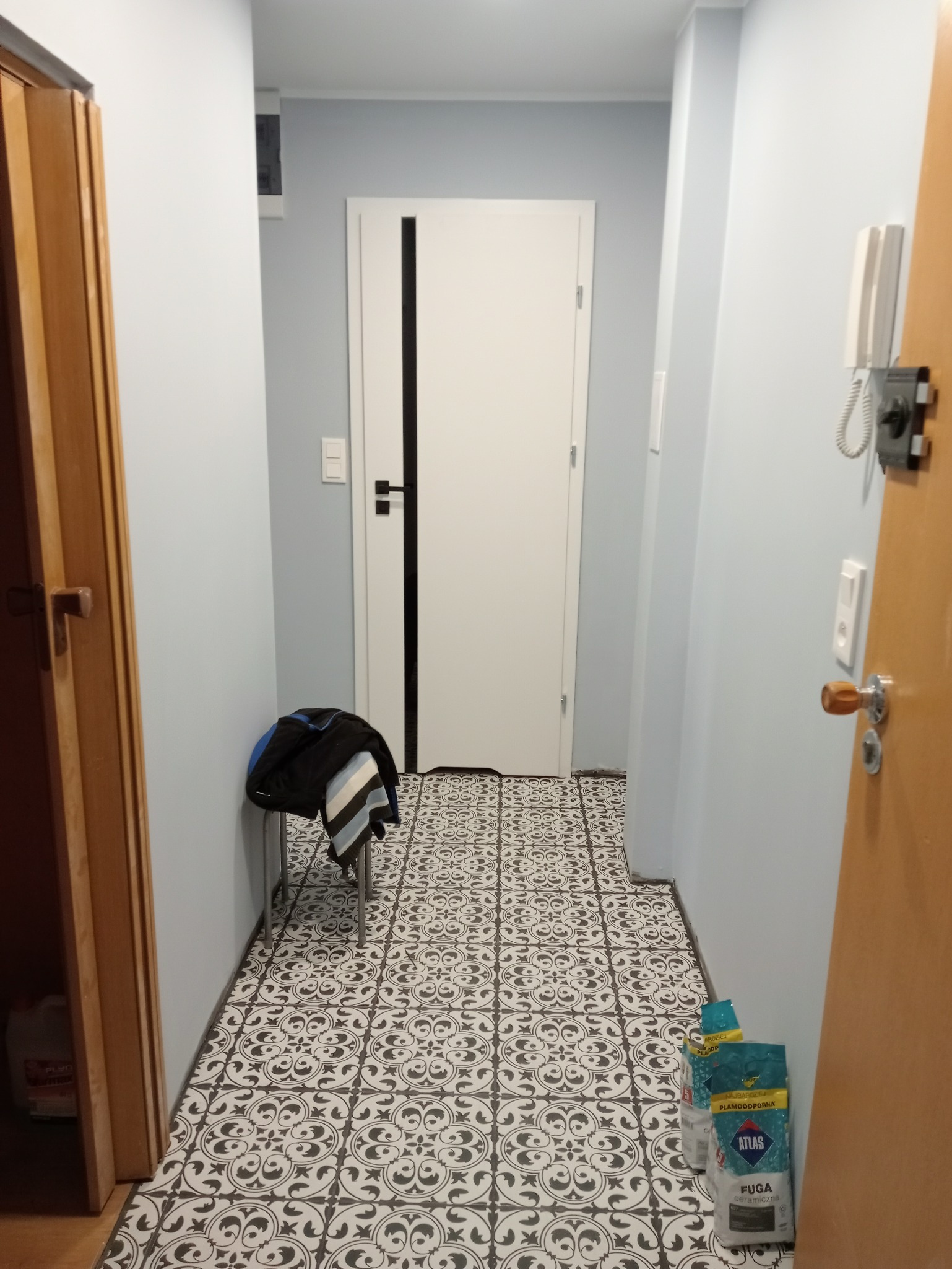 Korytarz po renowacji z wzorzystą, czarno-białą podłogą z płytek ceramicznych, białe drzwi z czarnym akcentem, jasnoniebieskie ściany, widoczne worki z fugą Atlas i krzesło z ubraniami.