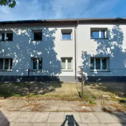 Dwukondygnacyjny budynek z białymi ścianami i szarą podmurówką, widoczny przez metalową siatkę ogrodzeniową, z cieniami drzew na elewacji i chodnikiem w pierwszym planie, cień osoby robiącej...