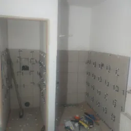 Łazienka w trakcie układania płytek ściennych, widoczne krzyżyki dystansowe, narzędzia i wiadro na podłodze, surowy stan, brak fug.