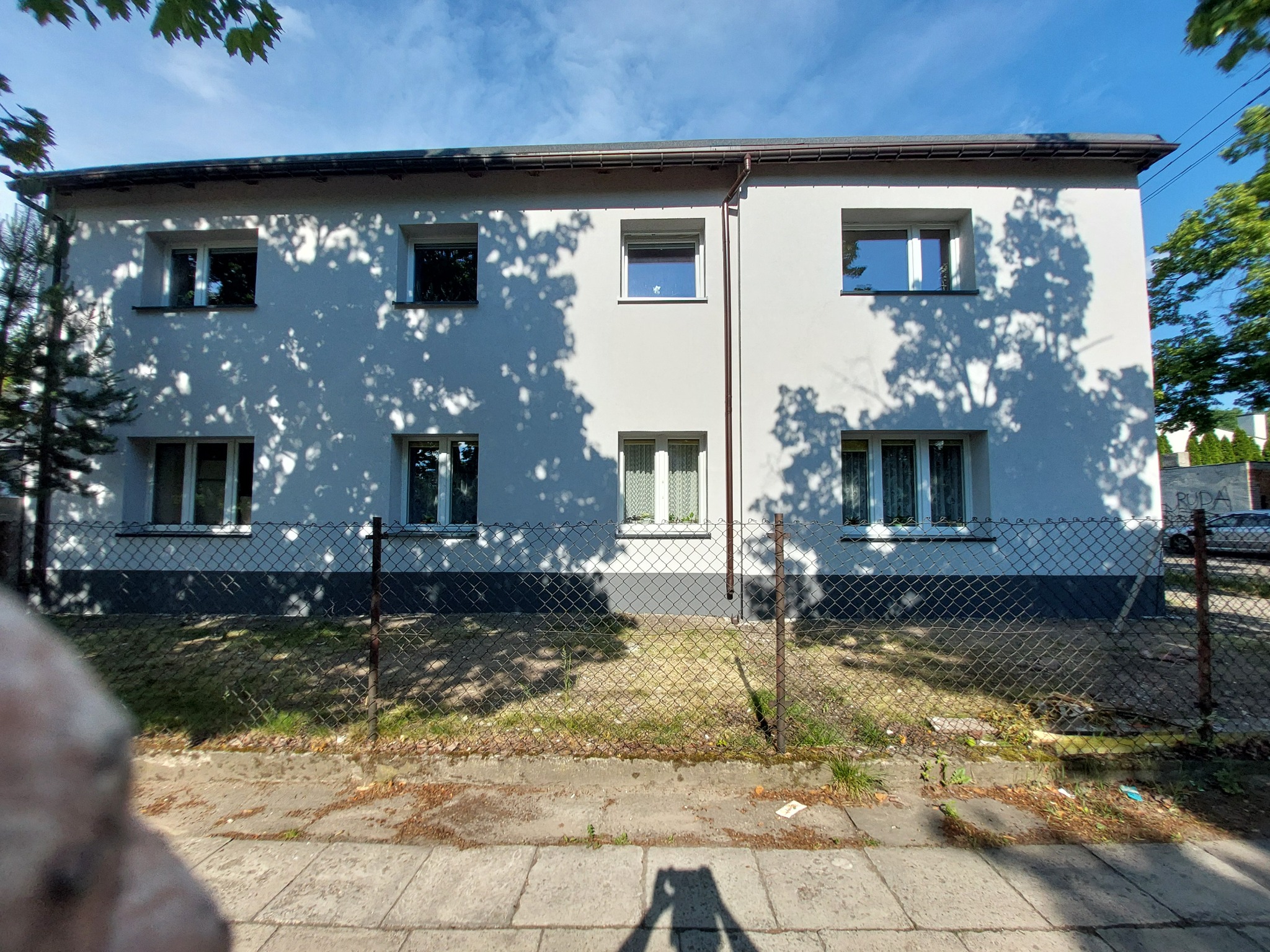 Dwukondygnacyjny budynek z białymi ścianami i szarą podmurówką, widoczny przez metalową siatkę ogrodzeniową, z cieniami drzew na elewacji i chodnikiem w pierwszym planie, cień osoby robiącej...