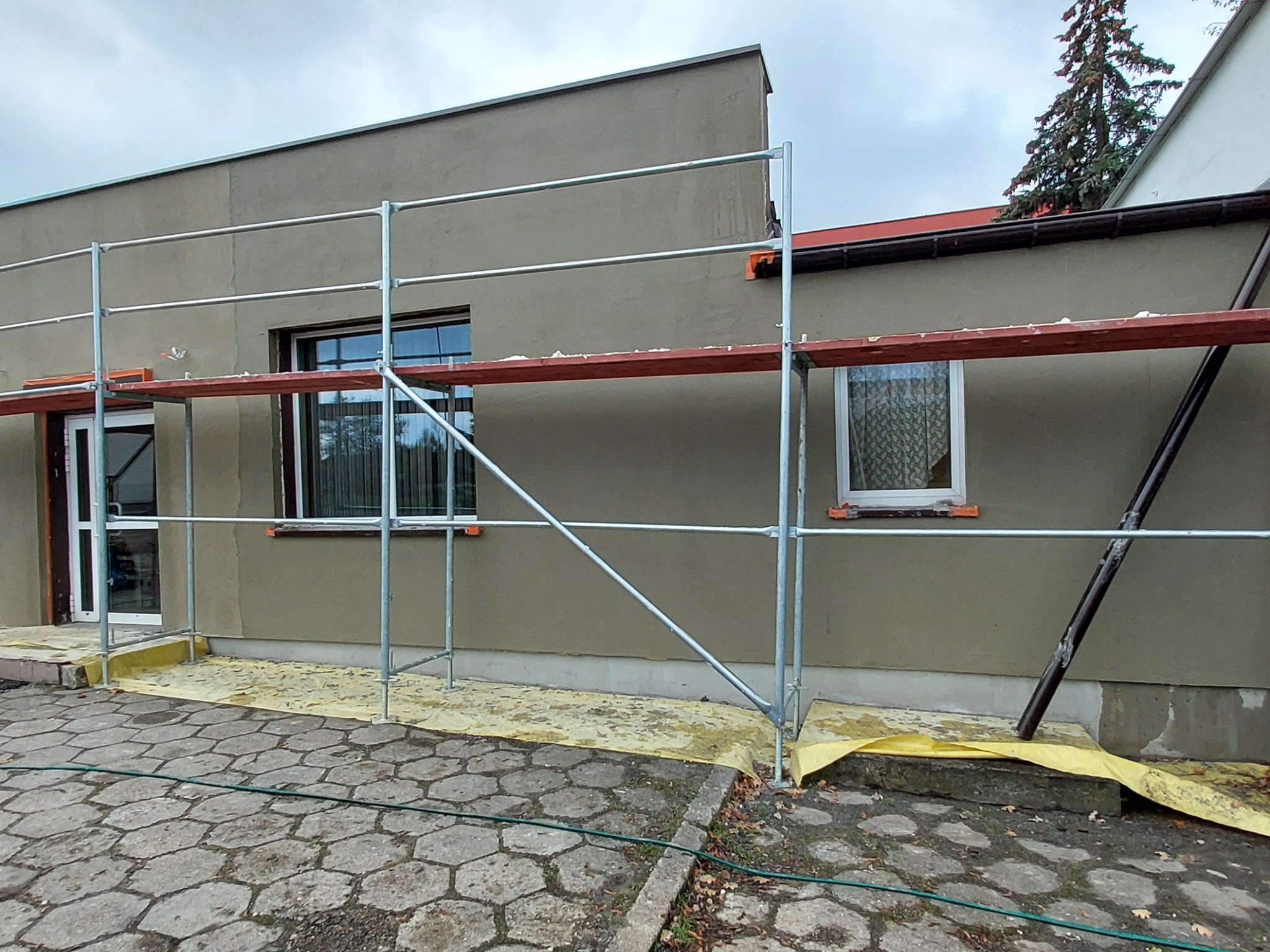 Budynek jednorodzinny w trakcie prac elewacyjnych, widoczne rusztowanie, zabezpieczona folią strefa przy budynku i świeżo nałożony tynk w kolorze beżowym.