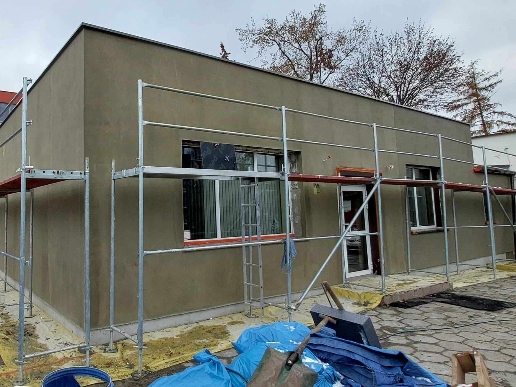 Budynek jednokondygnacyjny w trakcie prac elewacyjnych, z widocznym świeżo nałożonym tynkiem w odcieniach szarości, otoczony rusztowaniem. Na pierwszym planie narzędzia i materiały budowlane, w tle...