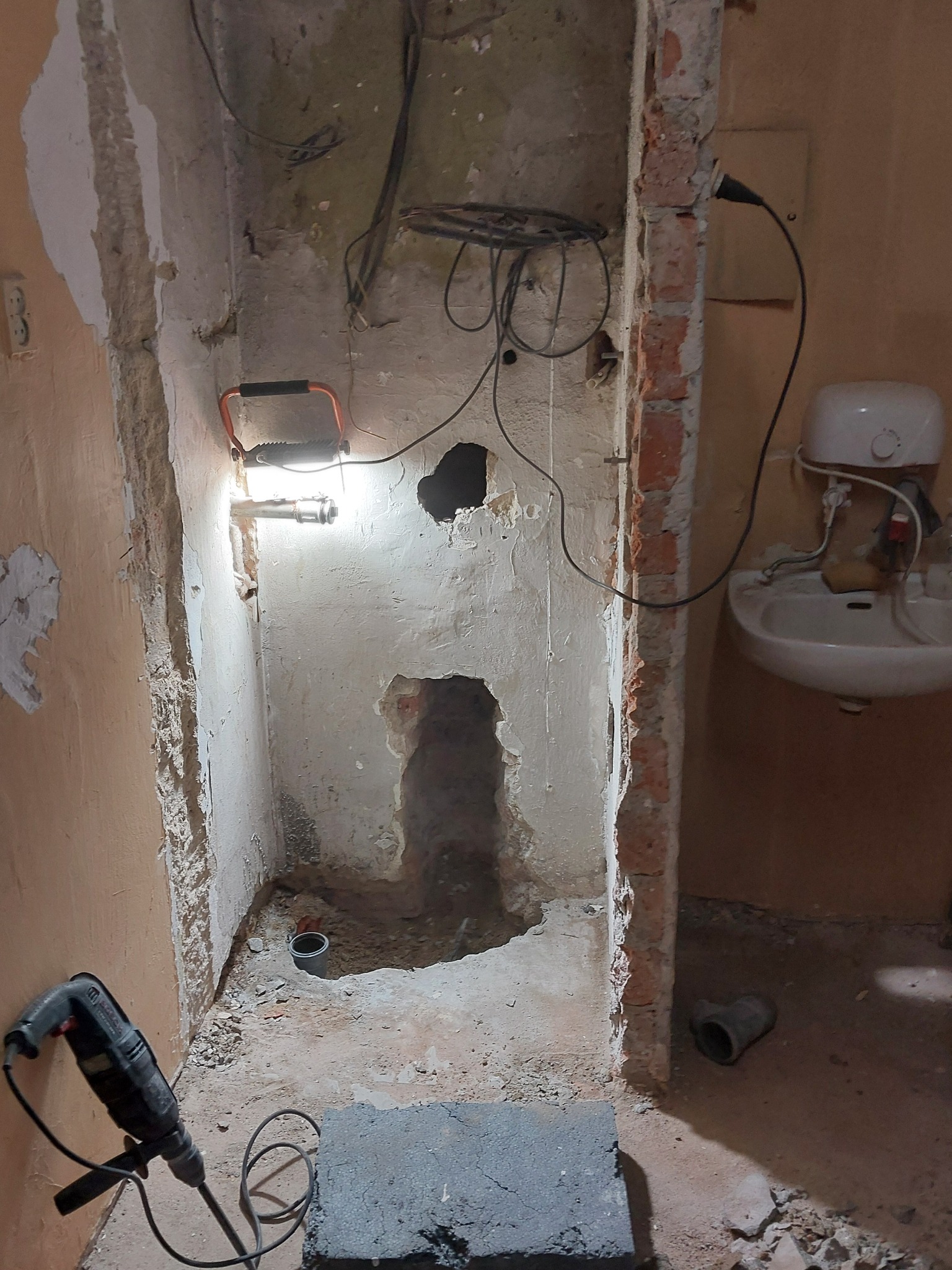 Rozkuta ściana w łazience podczas remontu, widoczny gruz, przewody, lampa robocza i wiertarka udarowa. Przygotowanie do instalacji hydraulicznej.