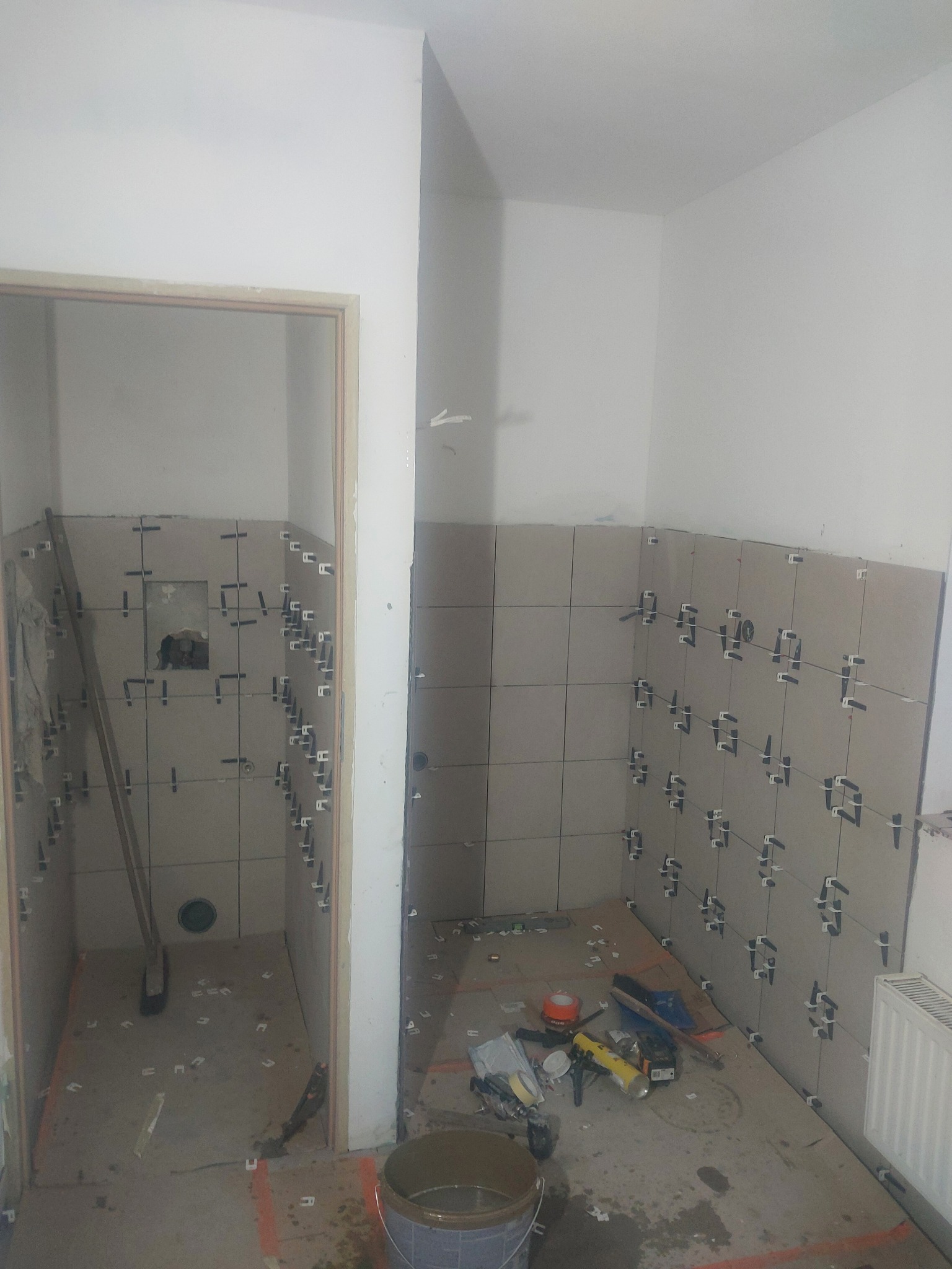 Łazienka w trakcie układania płytek ściennych, widoczne krzyżyki dystansowe, narzędzia i wiadro na podłodze, surowy stan, brak fug.