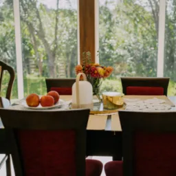 Stół z ciemnego drewna, nakryty koronkową serwetą, z bukietem astrów w wazonie, trzema jabłkami na talerzu i nowoczesną lampką, na tle okna z widokiem na ogród.