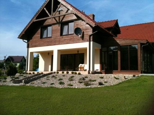 Dwukondygnacyjny dom z drewnianą elewacją, czerwoną dachówką i ogrodem z kamieniami oraz trawnikiem w słoneczny dzień.