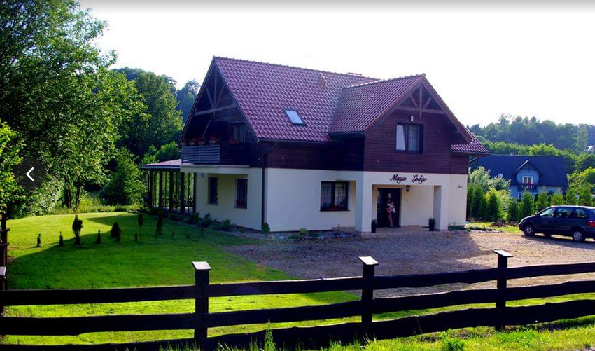 Dwupiętrowy budynek ośrodka terapii uzależnień z ciemnym drewnianym wykończeniem, jasną elewacją, czerwonym dachem i nazwą 'Maya Lodge' nad wejściem, otoczony zielonym trawnikiem i drewnianym...