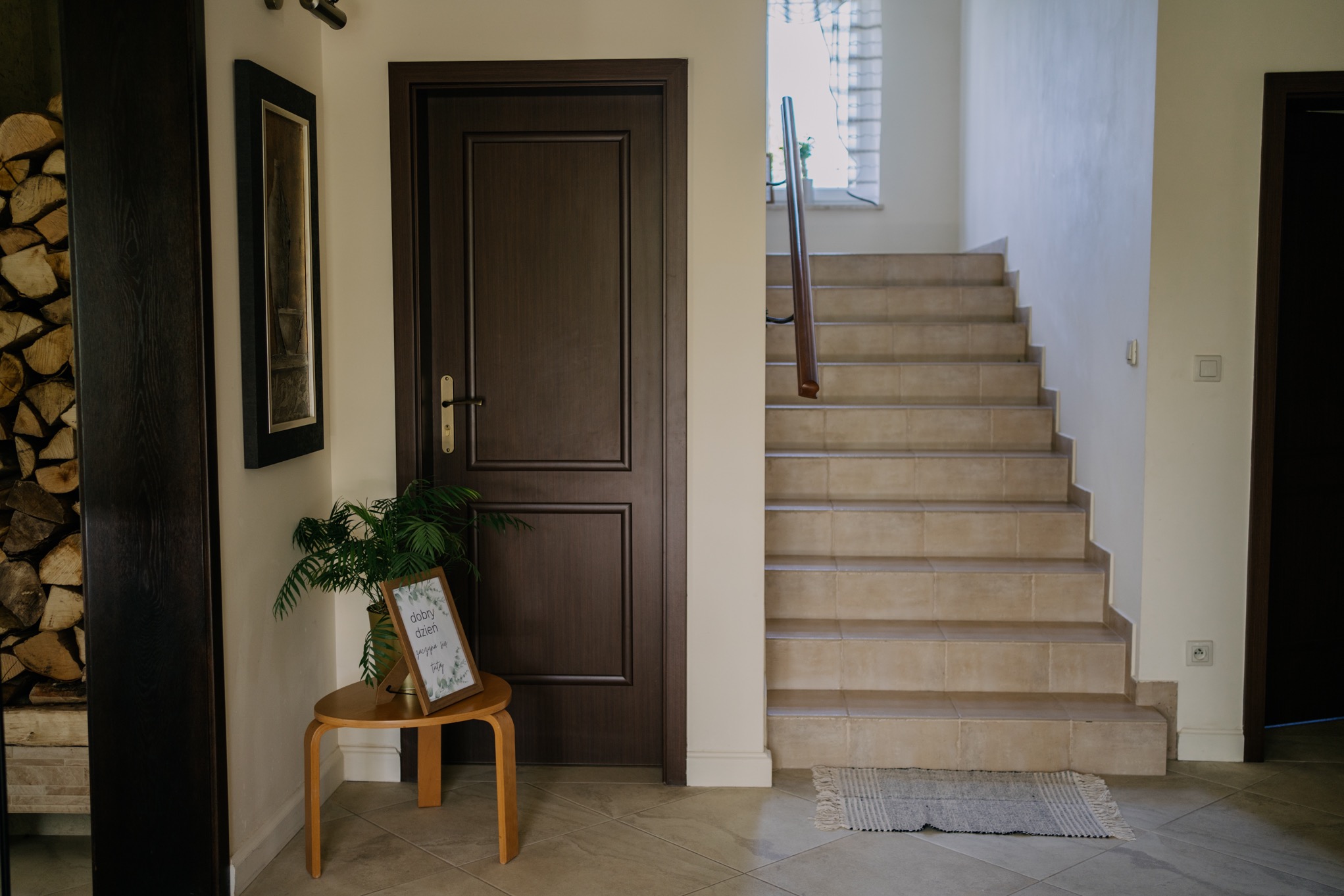 Wnętrze domu: drewniane drzwi, schody wyłożone beżowymi płytkami, dywanik, mały stolik z rośliną doniczkową i ramką z napisem 'Dobry dzień zaczyna się tutaj'.