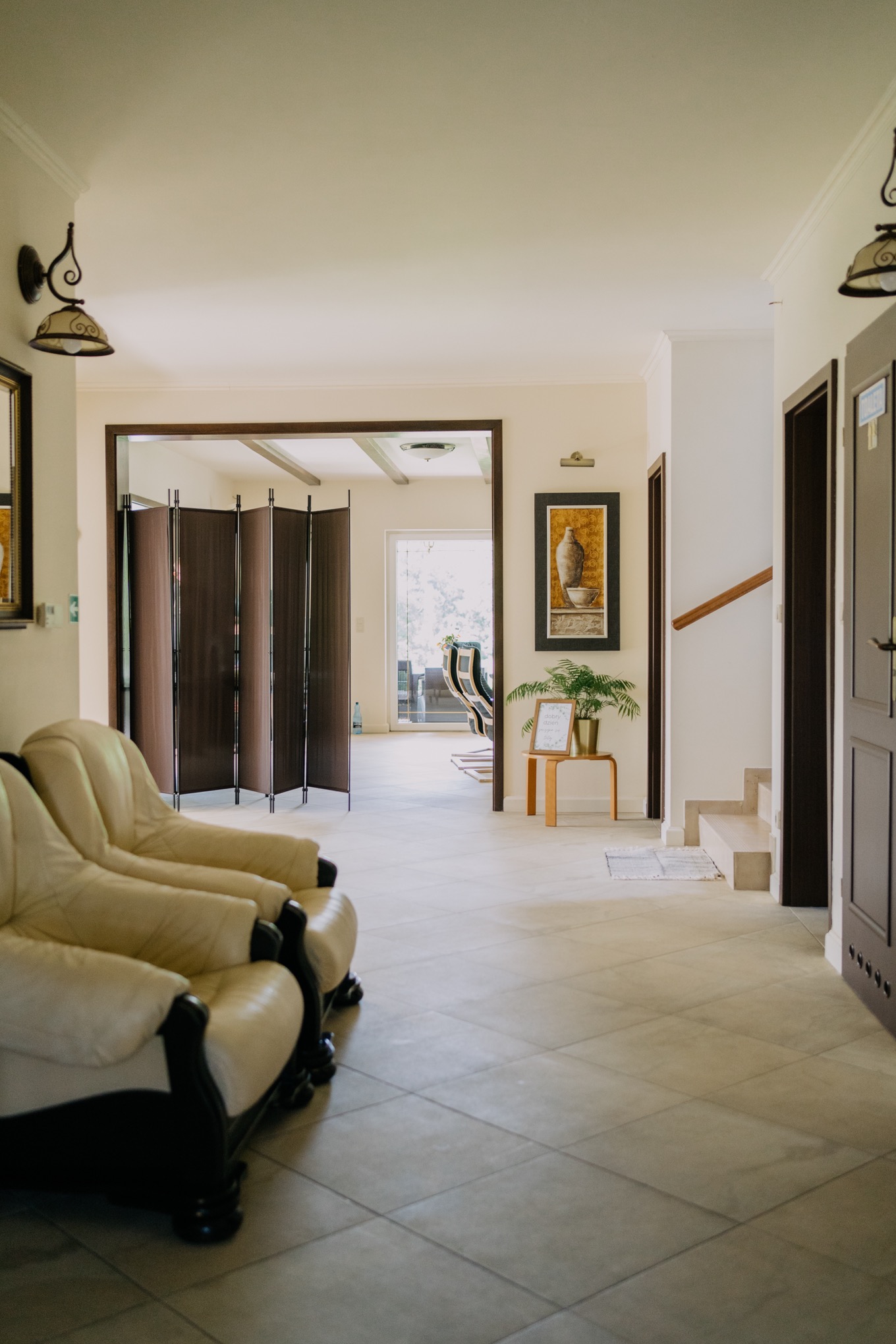 Jasne wnętrze z kremowymi fotelami, parawanem, obrazem i schodami, tworzące spokojną atmosferę poczekalni lub miejsca relaksu.