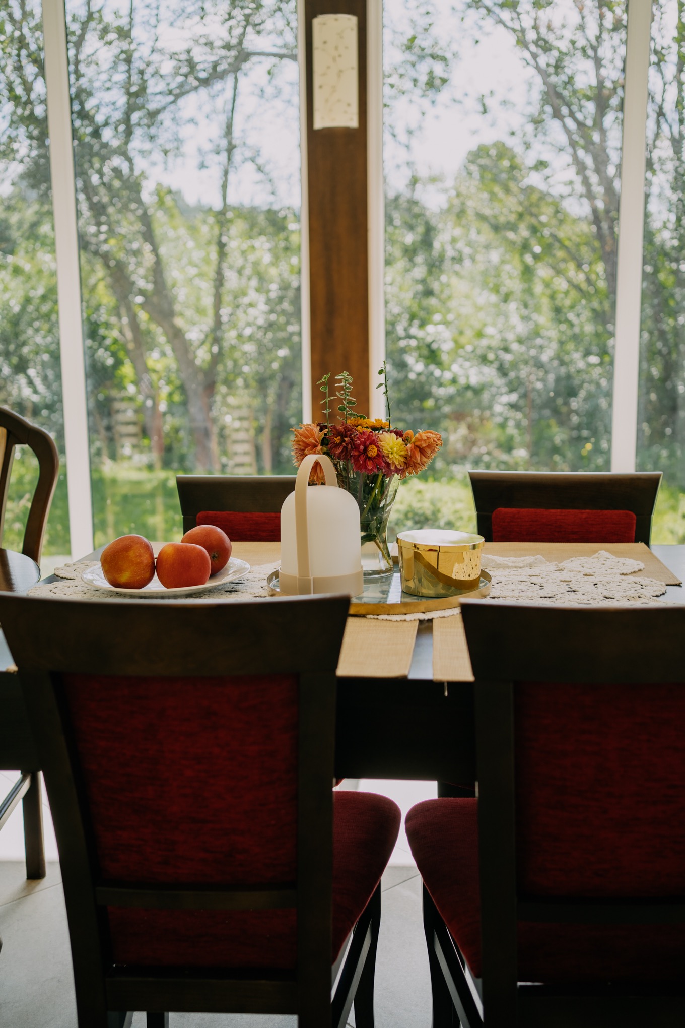 Stół z ciemnego drewna, nakryty koronkową serwetą, z bukietem astrów w wazonie, trzema jabłkami na talerzu i nowoczesną lampką, na tle okna z widokiem na ogród.