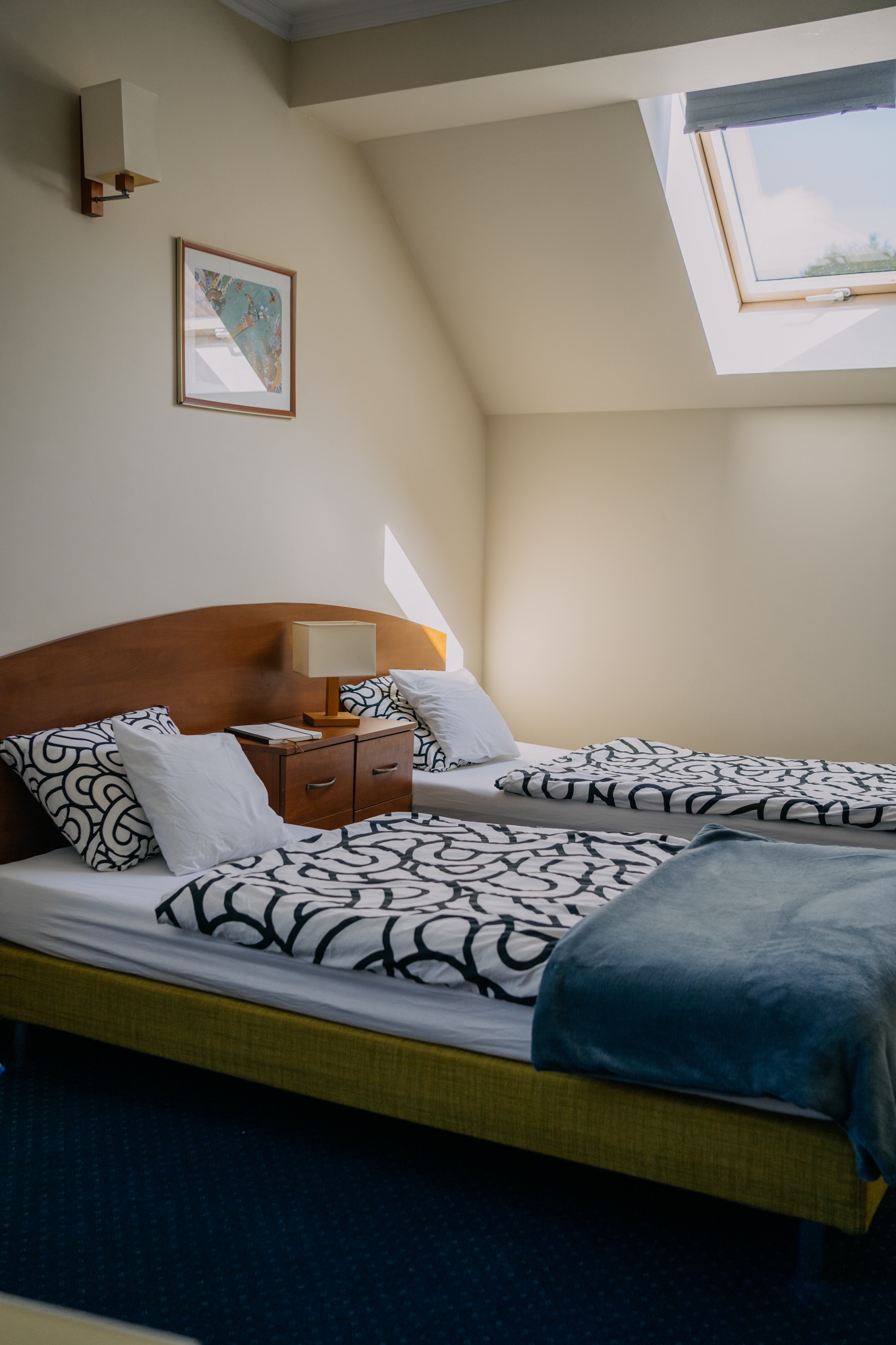 Pokój z dwoma łóżkami w jasnych kolorach, z drewnianym zagłówkiem, wzorzystą pościelą w czarno-białe wzory, nocną szafką z lampką i notatnikiem, obrazkiem na ścianie i oknem dachowym.