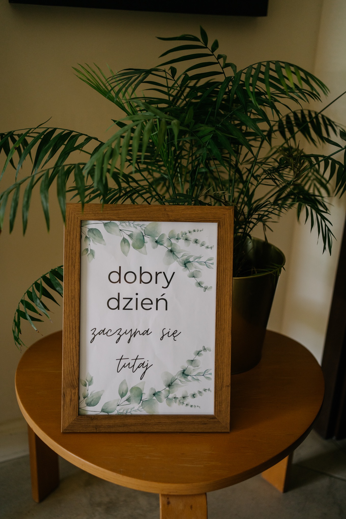 Oprawiona grafika z napisem 'dobry dzień zaczyna się tutaj' z motywem roślinnym, umieszczona na okrągłym stoliku obok rośliny doniczkowej.