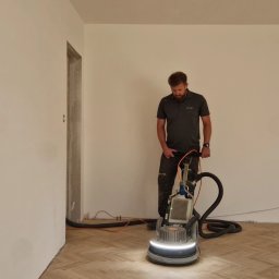 Marcin Ciepły Floorlab Podłogi Drewniane - Mężczyzna obsługuje maszynę do obróbki parkietu ułożonego we wzór jodełki francuskiej, widoczne ślady obróbki, w tle surowe ściany i widoczna instalacja elektryczna.