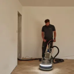 Mężczyzna obsługuje maszynę do obróbki parkietu ułożonego we wzór jodełki francuskiej, widoczne ślady obróbki, w tle surowe ściany i widoczna instalacja elektryczna.