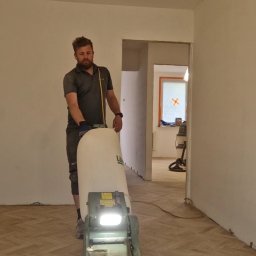 Marcin Ciepły Floorlab Podłogi Drewniane - Mężczyzna z brodą cyklinuje parkiet jodełkowy w jasnym pomieszczeniu za pomocą profesjonalnej maszyny z oświetleniem i workiem na pył.