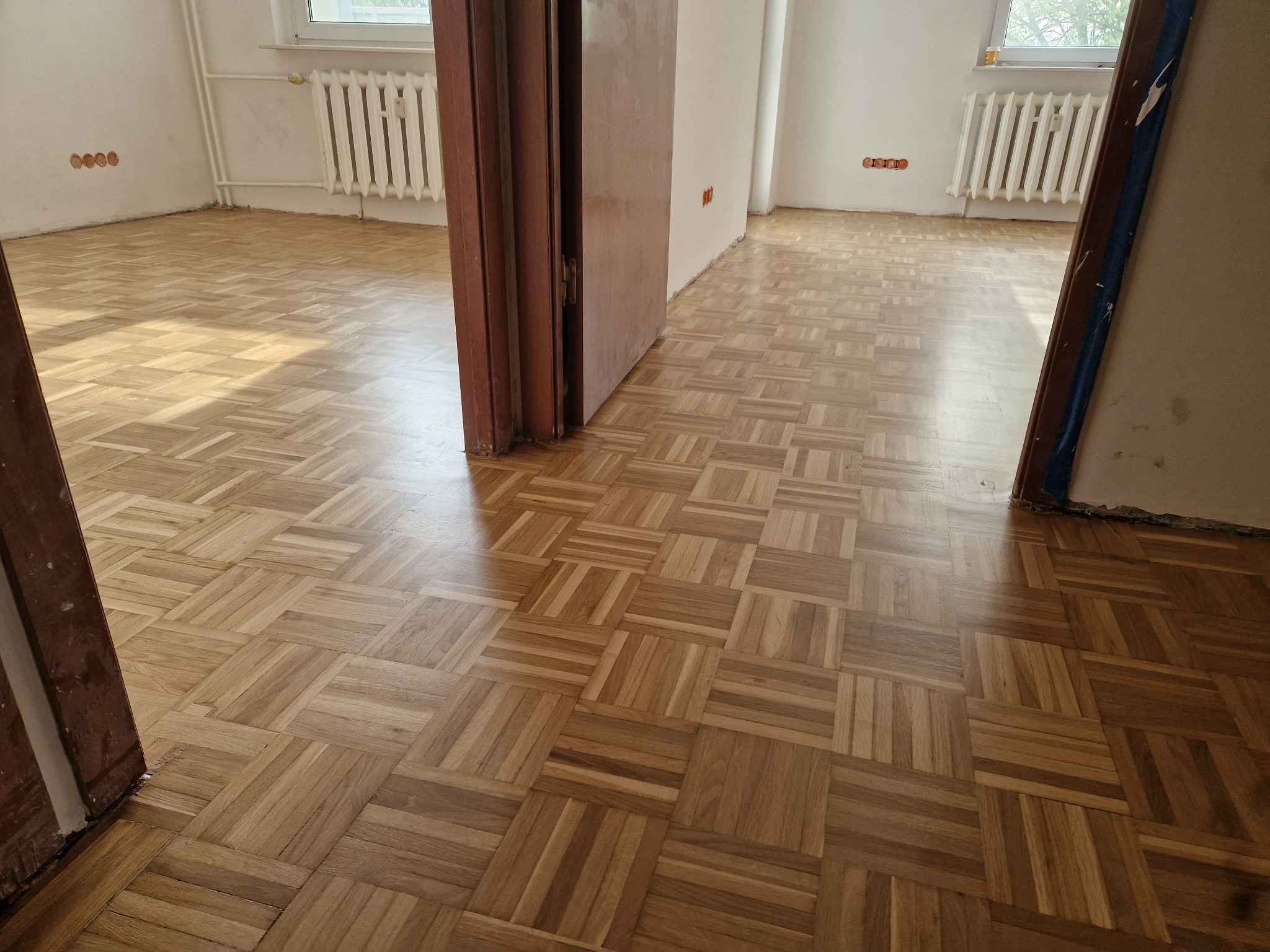 Ułożony parkiet w jodełkę francuską w pustym pokoju z otwartymi drzwiami, widoczne grzejniki i ślady po gniazdkach elektrycznych na ścianach.