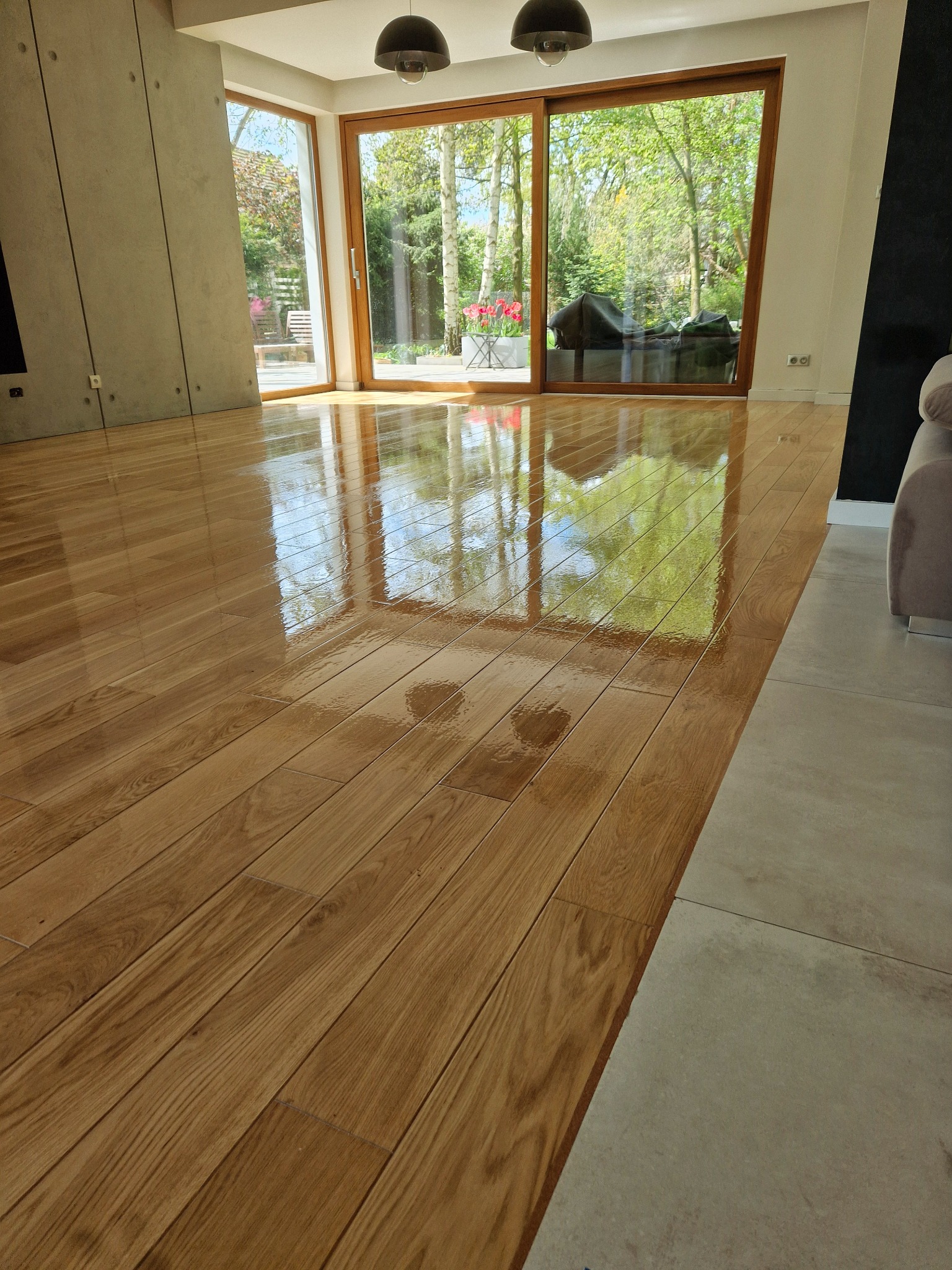 Błyszcząca, świeżo wykończona drewniana podłoga w jasnym salonie, z widocznym odbiciem okna i ogrodu, sąsiadująca z szarą, matową płytką podłogową.