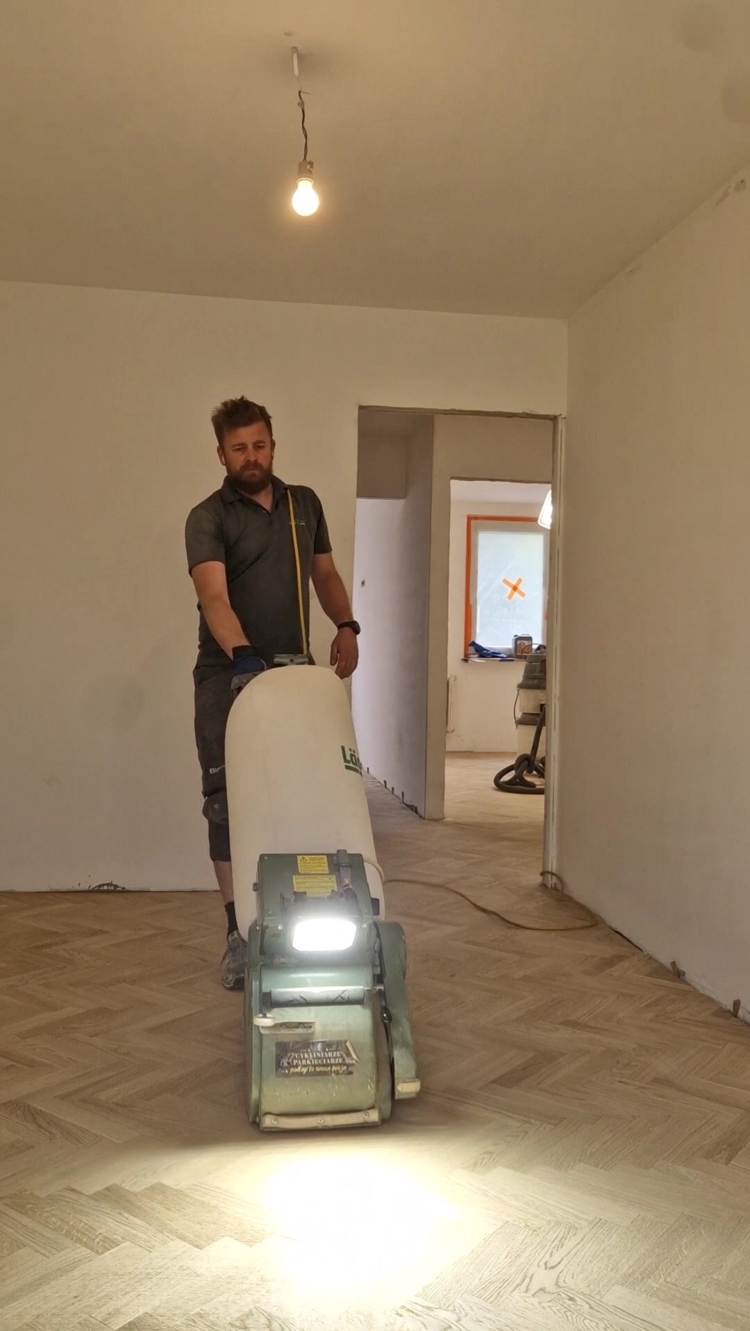 Mężczyzna z brodą cyklinuje parkiet jodełkowy w jasnym pomieszczeniu za pomocą profesjonalnej maszyny z oświetleniem i workiem na pył.