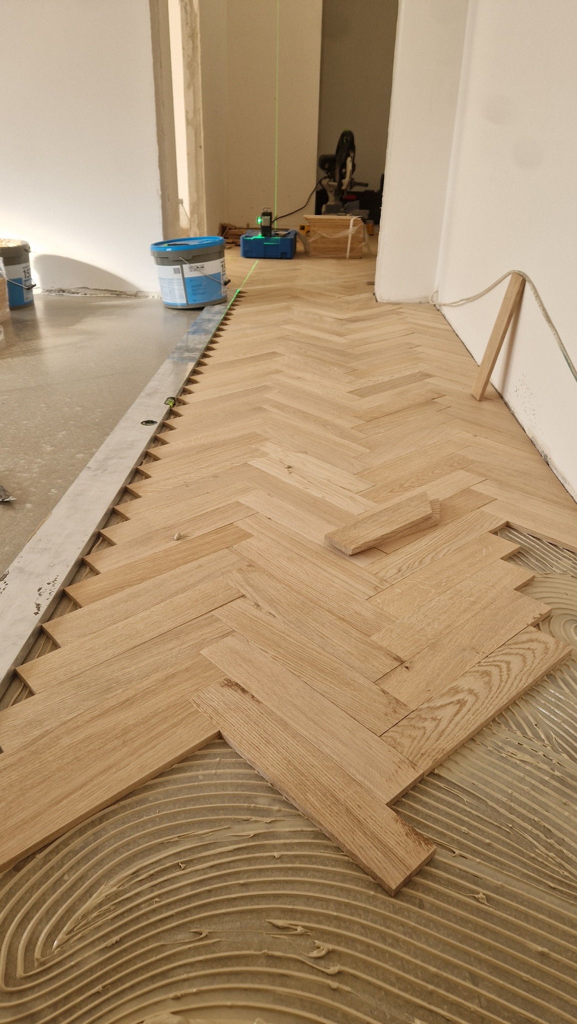 Układanie parkietu w jodełkę: widok z góry na klej rozprowadzony w łuki, deski parkietowe w trakcie montażu, poziomica laserowa i narzędzia w tle.